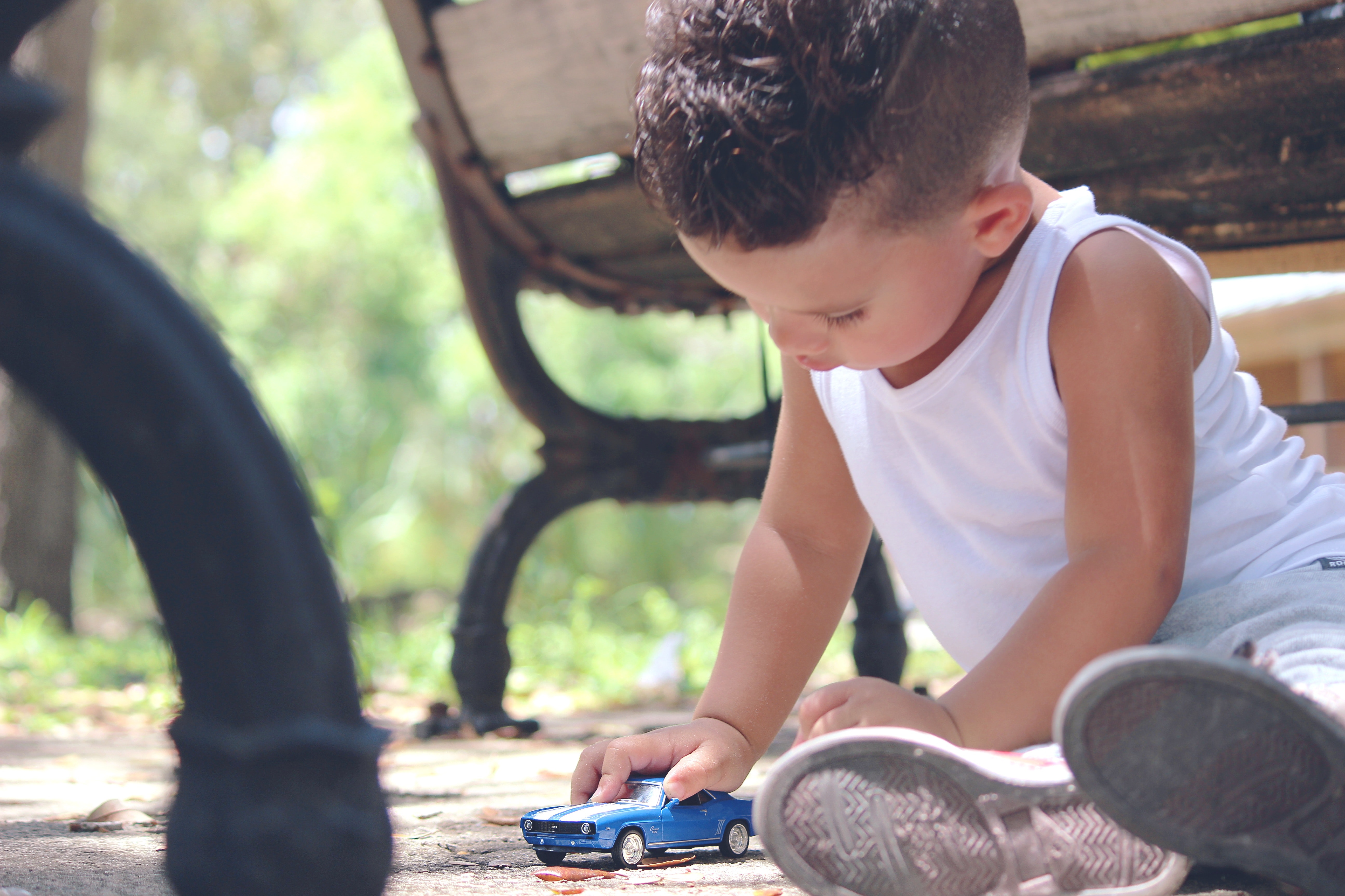 Boy in white tank top playing blue coupe die cast near brown wooden bench chair during daytime photo