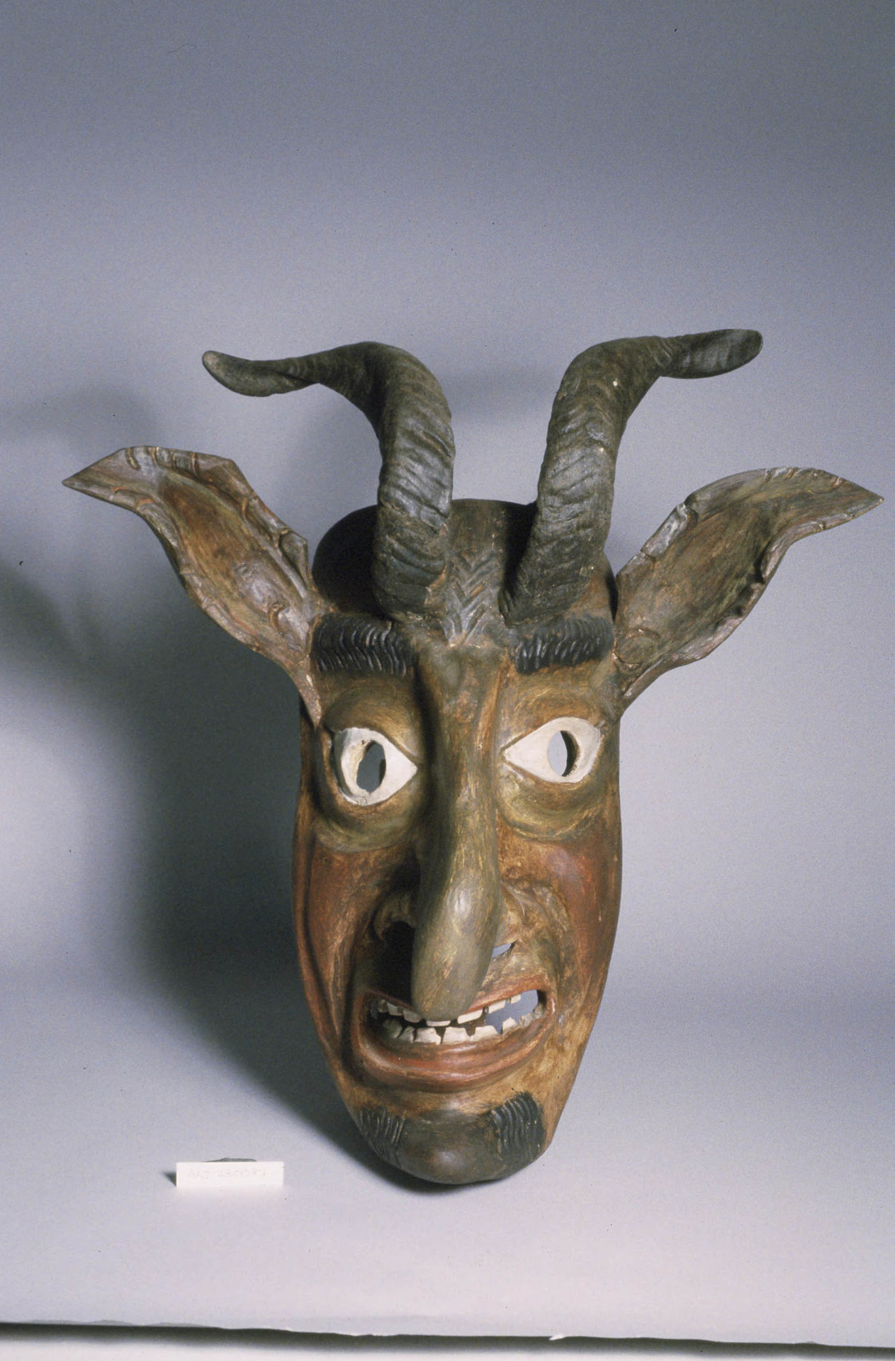 Carved wooden mask resembling an anthropomorphic goat. Mask has a ...
