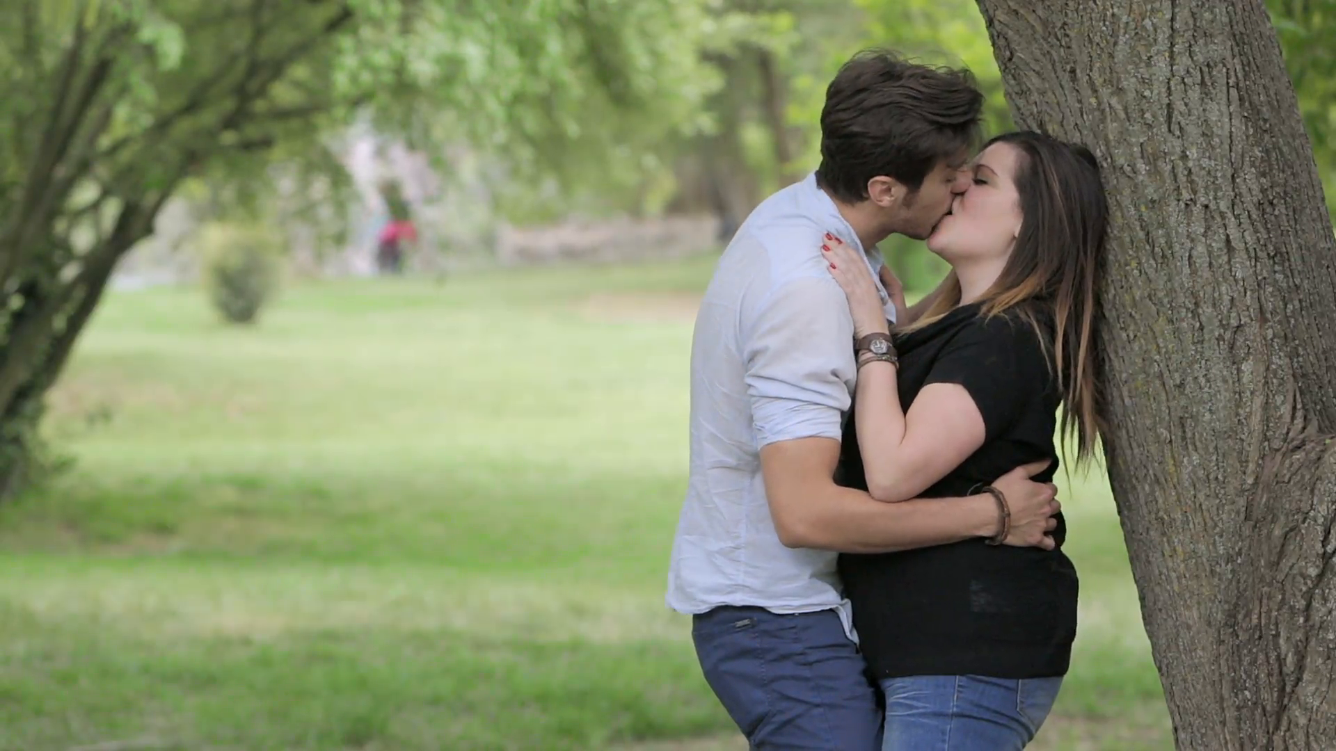 two young people kissing in a public park under a tree: couple in ...