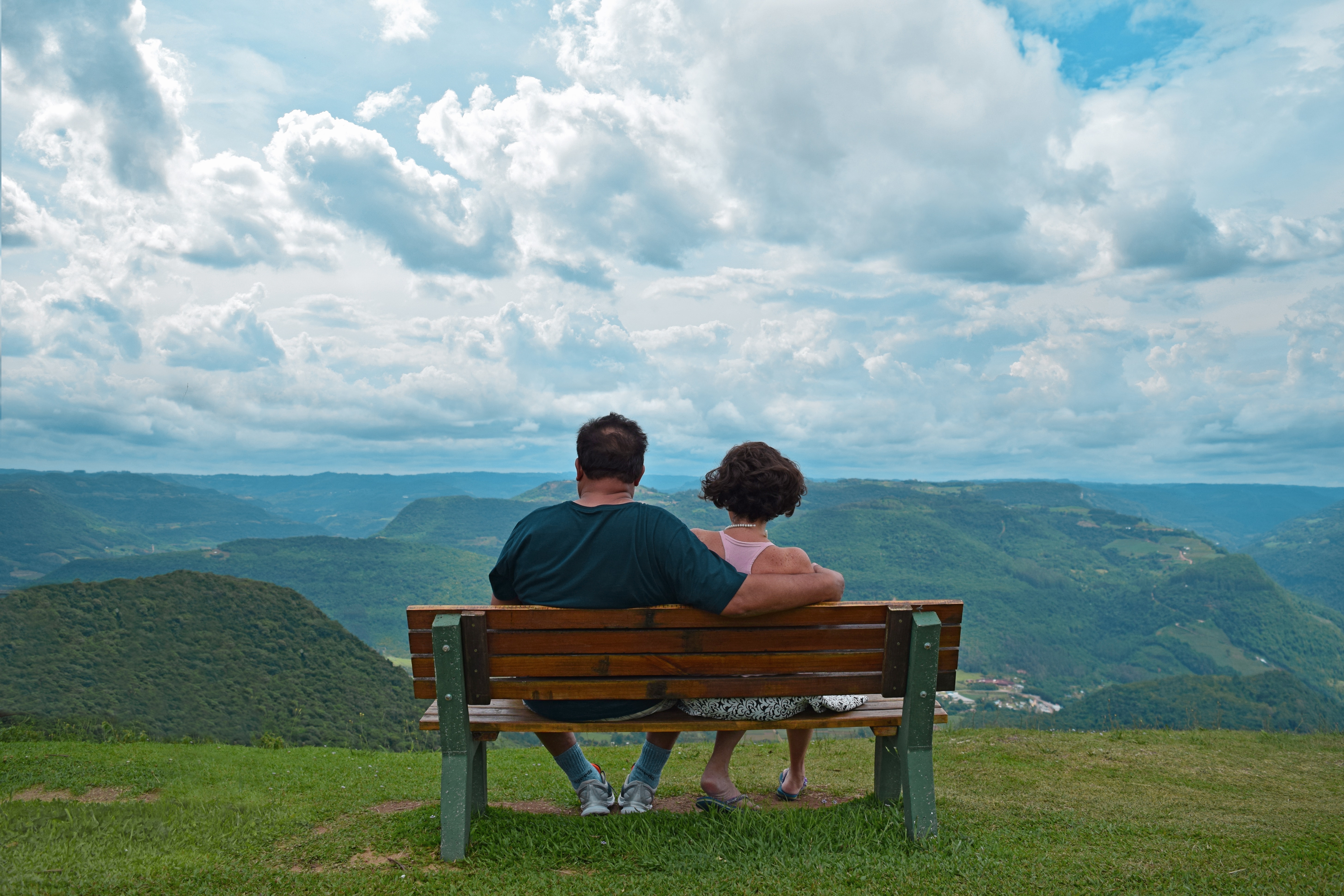 Couple sitting on brown wooden bench near mountains covered with grasses under blue cloudy sky photo