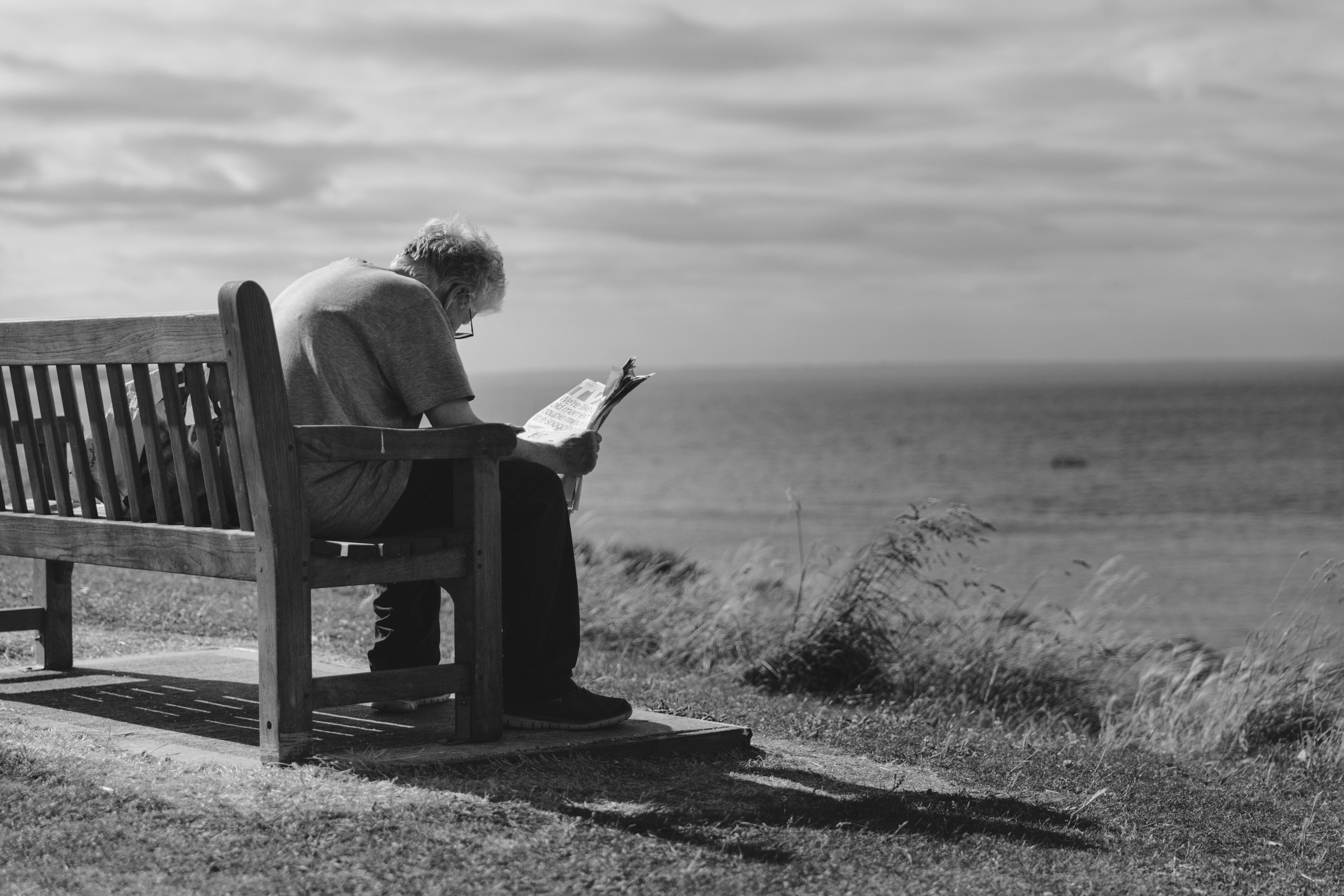 Grayscale Photo of Man Sitting on Brown Wooden Bench Reading News Paper during Day Time, Adult, Old man, Seaside, Seashore, HQ Photo