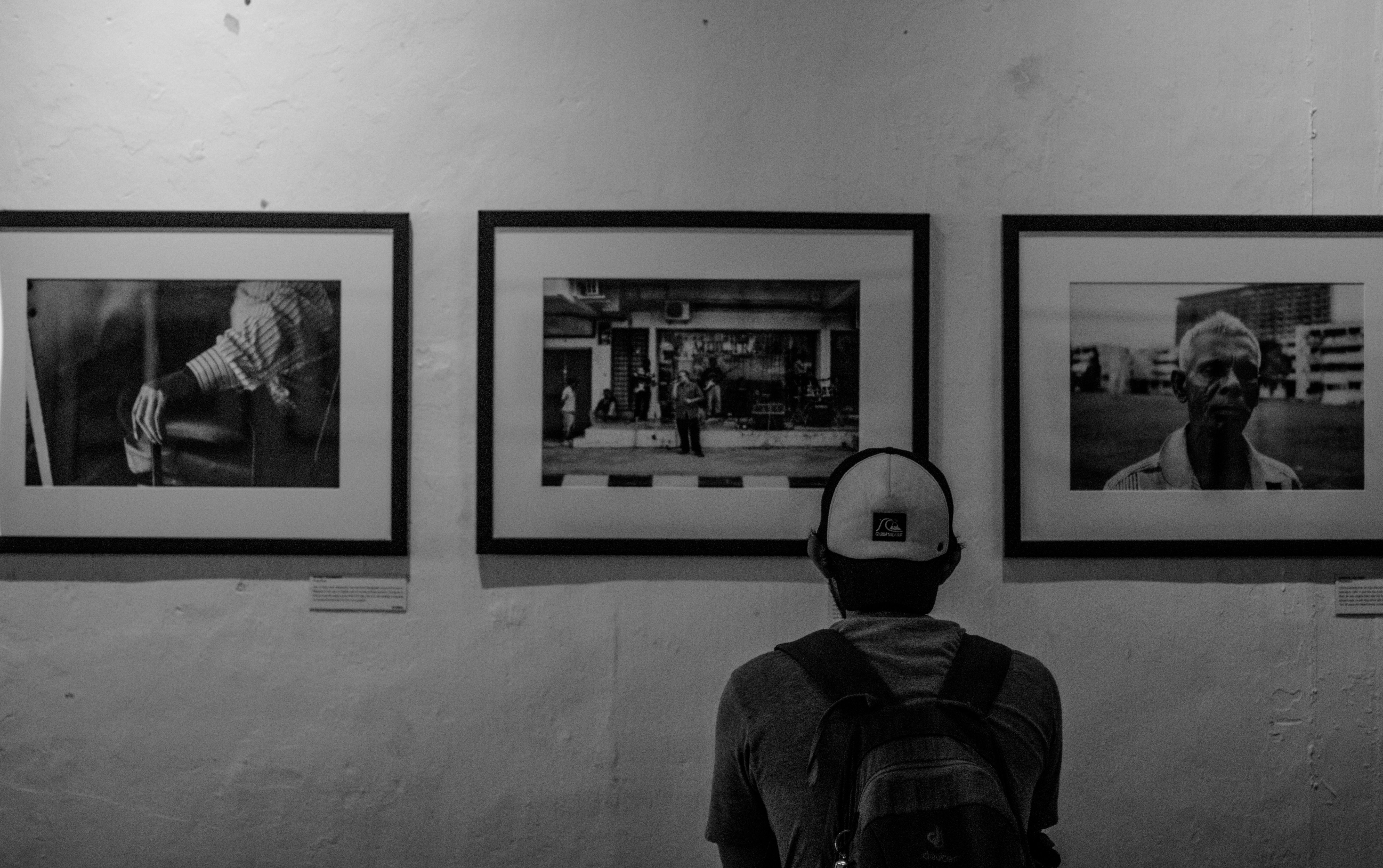 Grayscale photo of man wearing white cap in front of three paintings