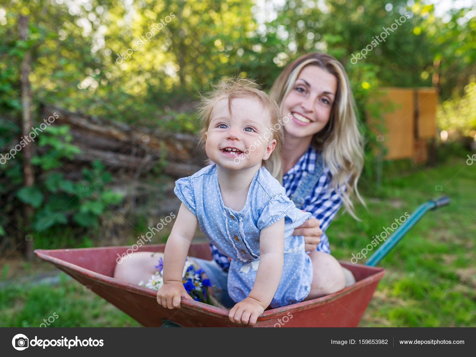 Mother and her little daughter sit in the wheelbarrow outdoors ...