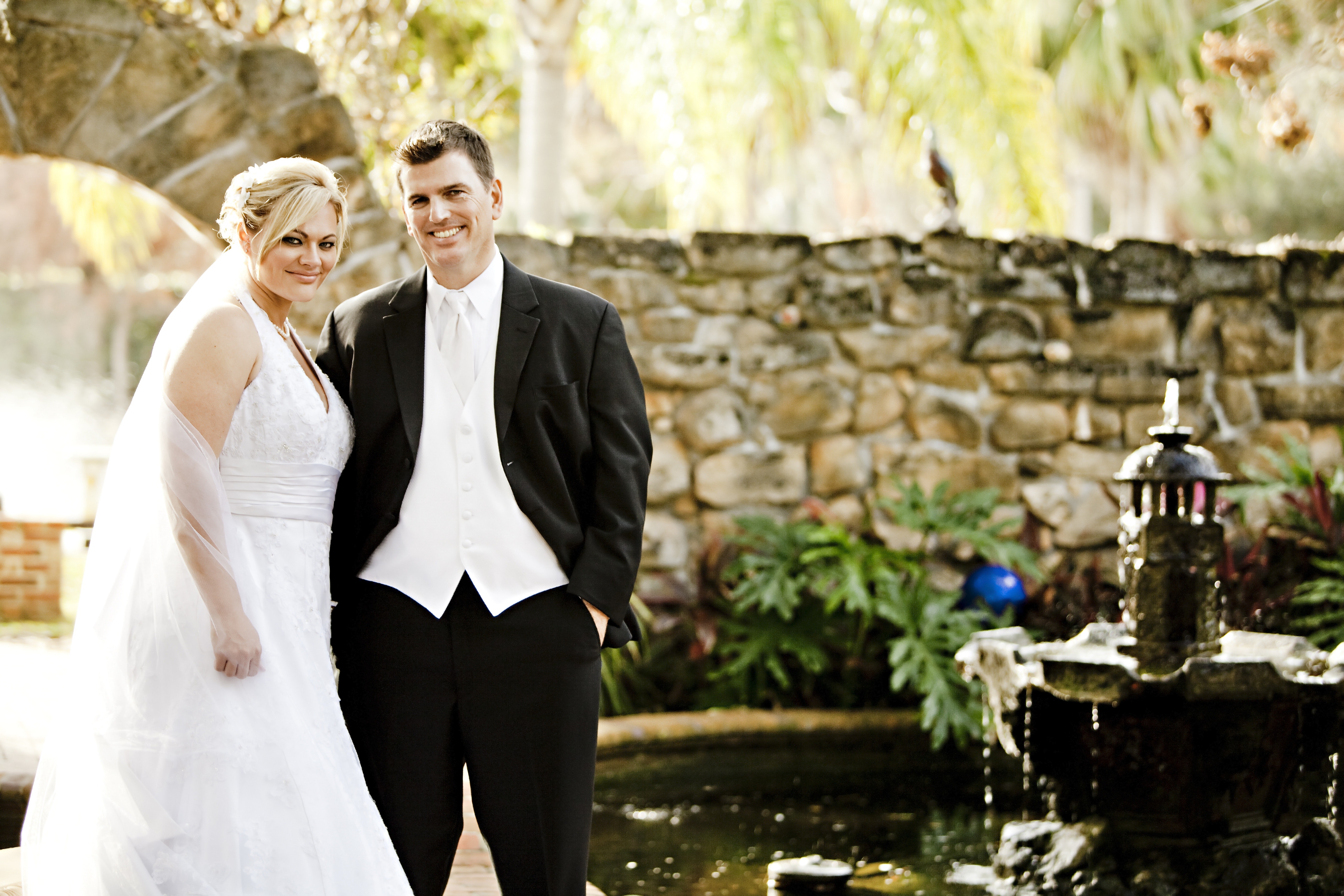 Man and woman in their wedding outfit with brown wall in the background near fountain and pond during daytime photo