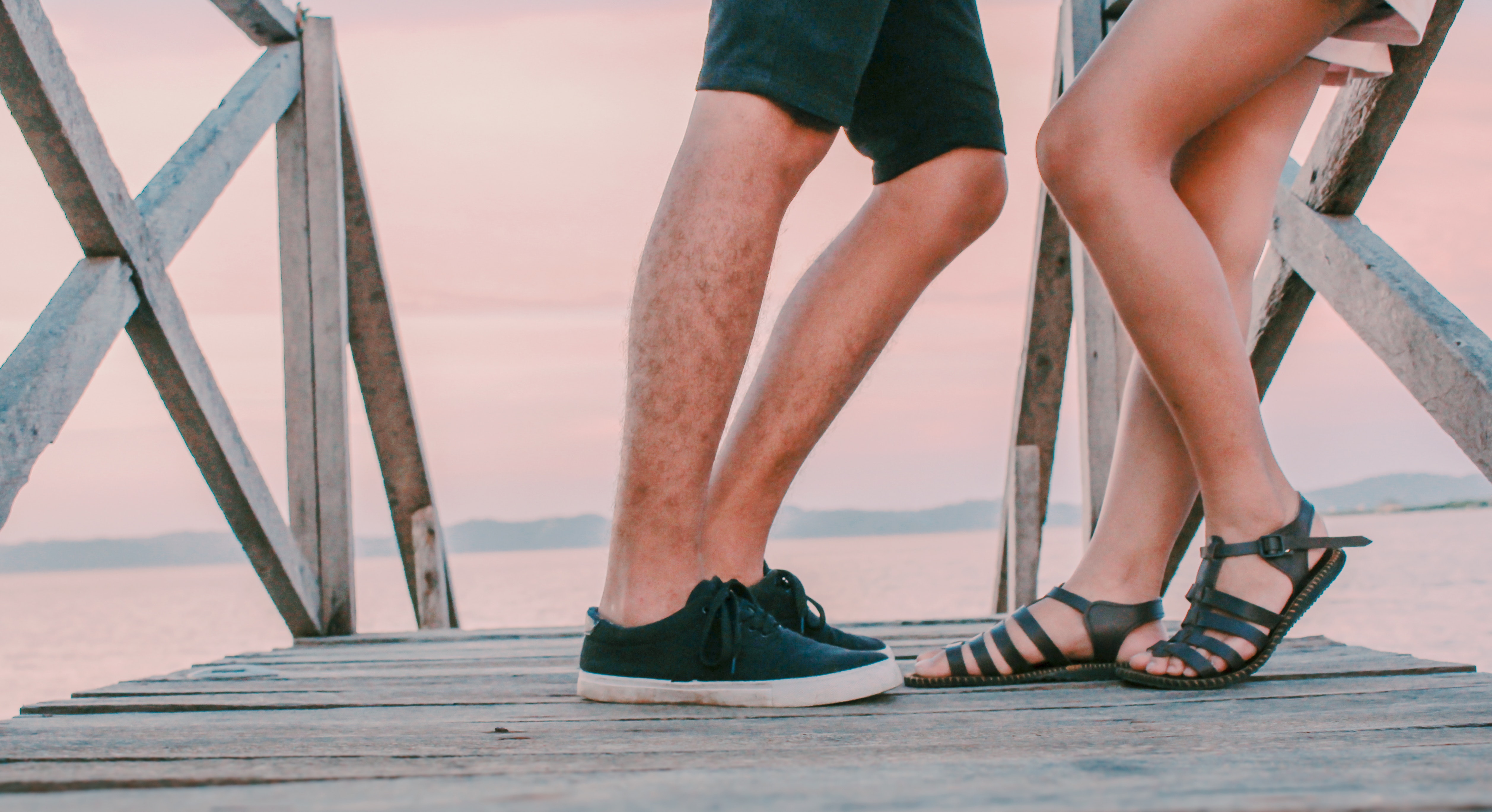 Man in black low-top sneakers leaning towards woman in black gladiator flat sandals on dock photo