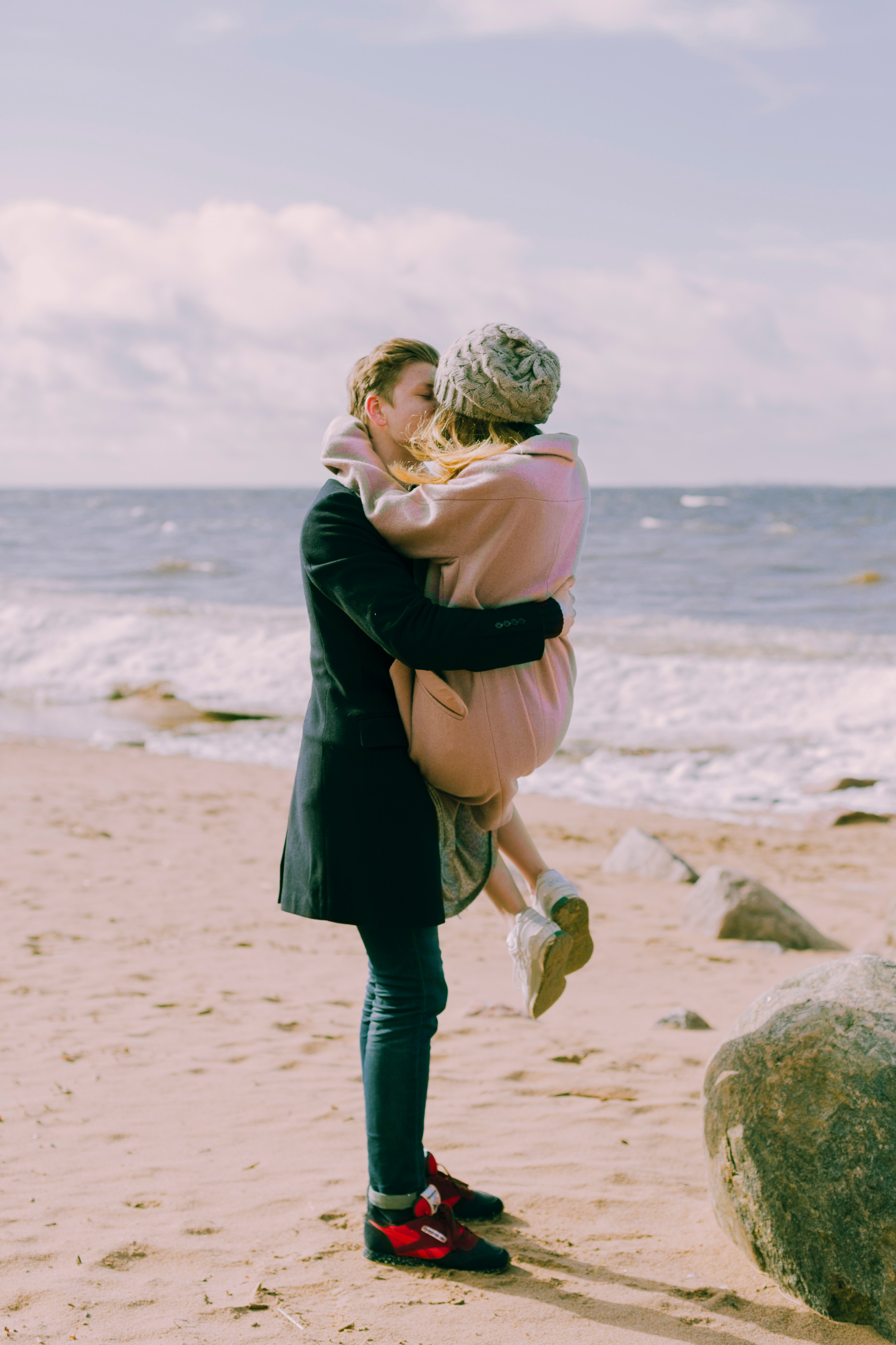 Man in black overcoat and blue denim jeans kissing while carrying a woman in pink overcoat and knit cap on shore at daytime photo