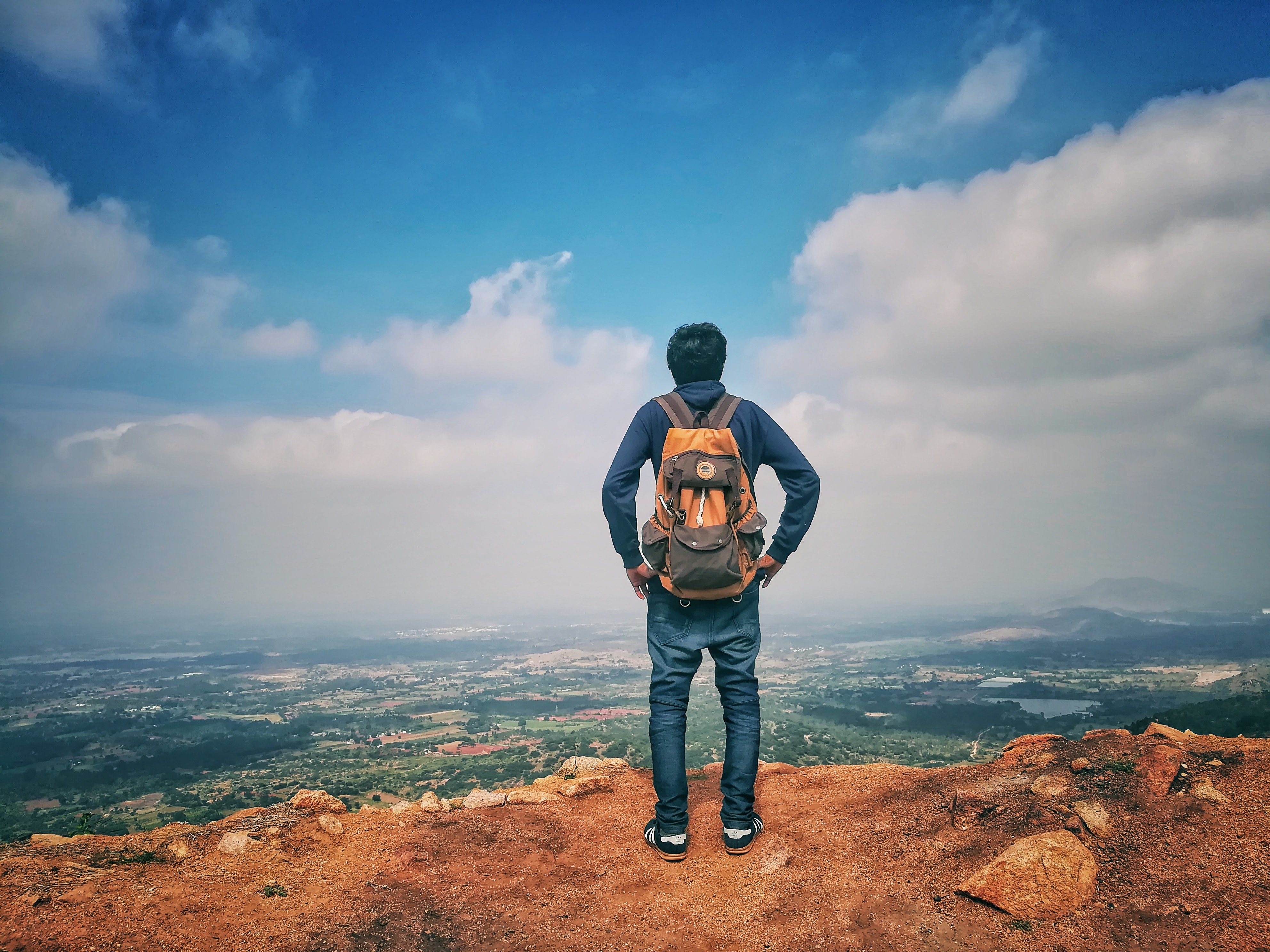 Man in blue dress shirt and blue jeans and orange backpack standing on mountain cliff looking at town under blue sky and white clouds photo