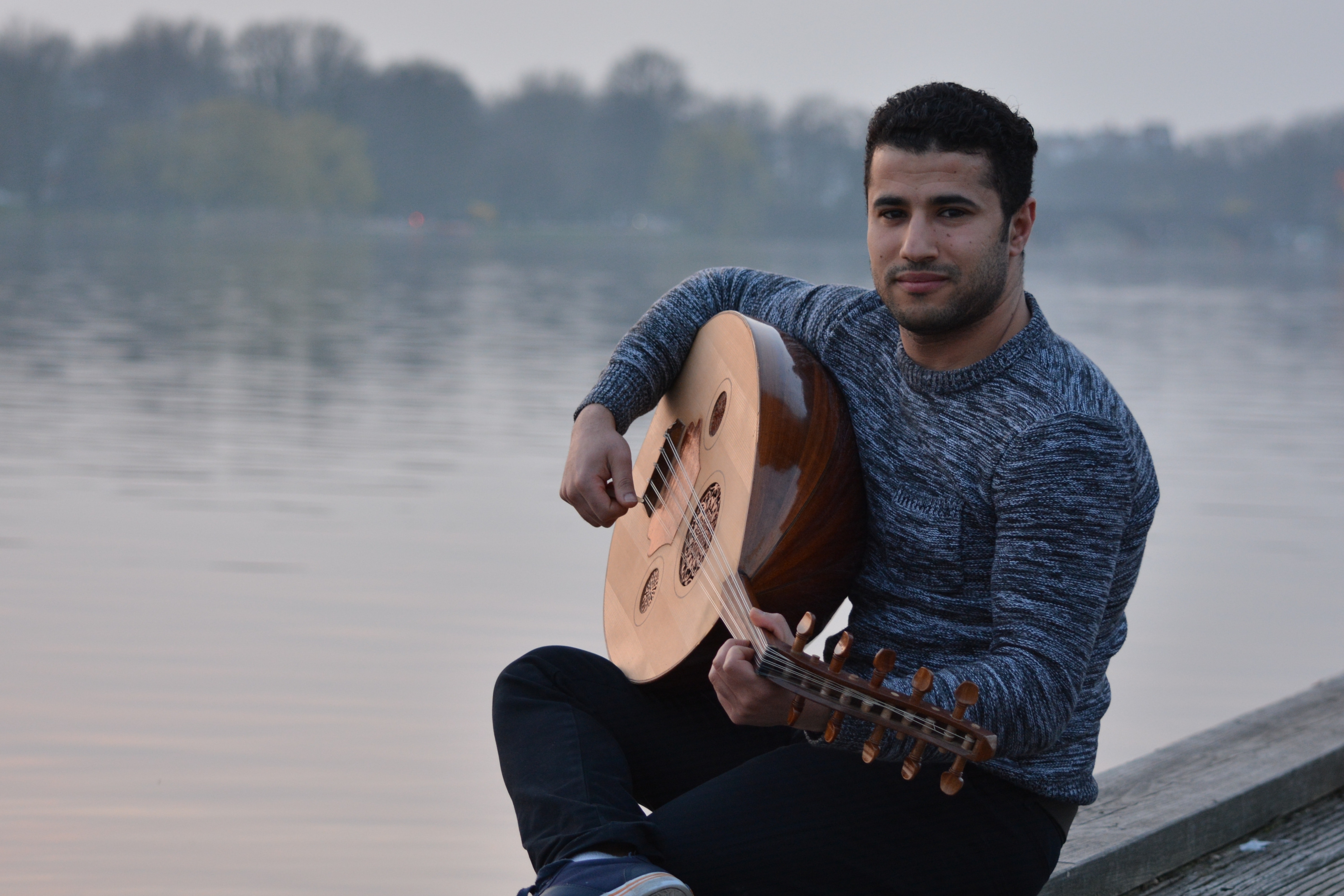 Man in gray crew neck sweatshirt and black pants sitting in gray concrete holding string instrument near body of water photo