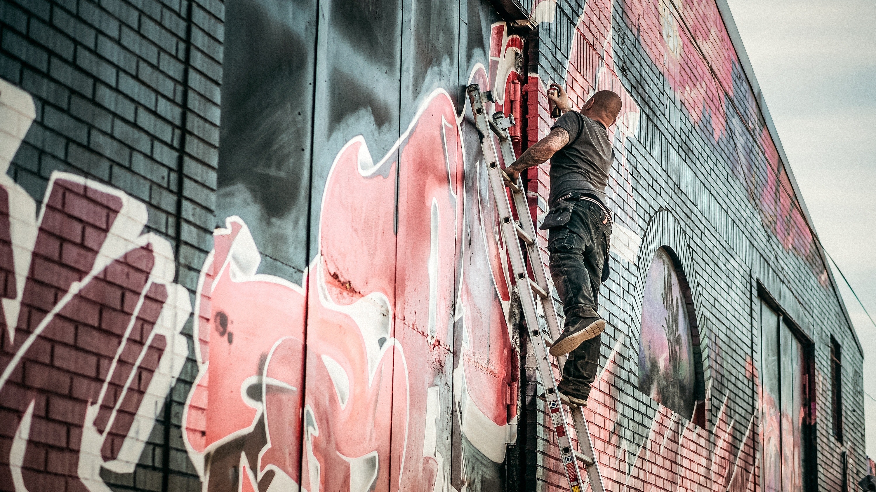 Man in gray shirt standing on gray steel ladder painting black white and red graffiti on concrete wall outdoors photo