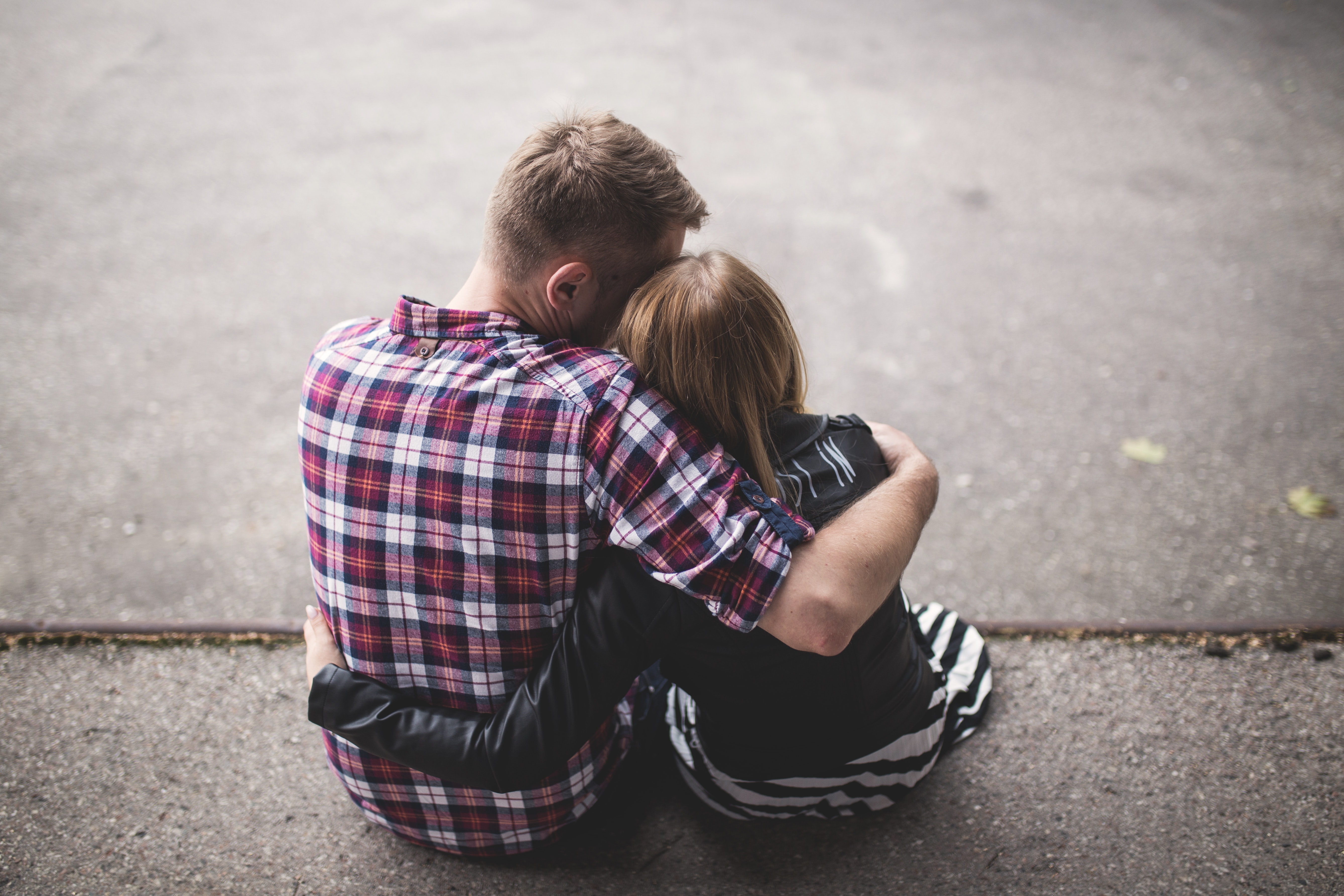 Man in red white and blue check long sleeve shirt beside woman in black and white stripes shirt hugging each other while sitting on a concrete surface photo