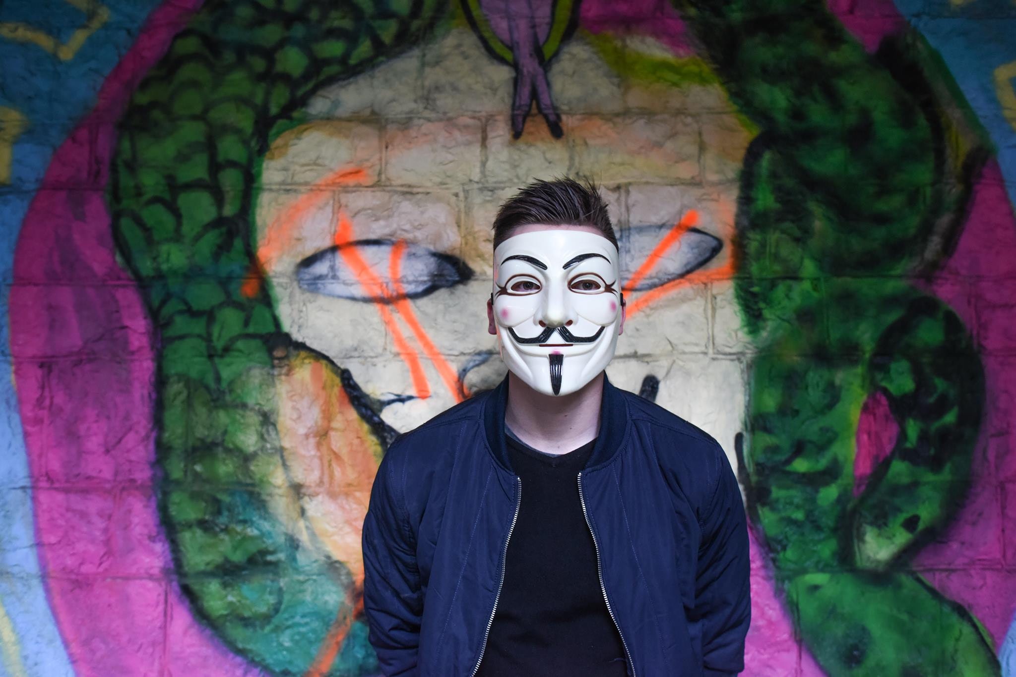 Man in white mask in black crew neck shirt and blue zip up jacket infront graffiti wall photo