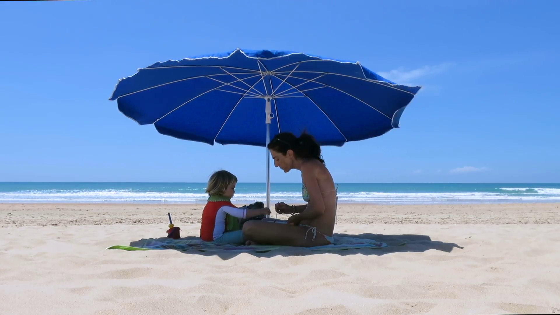 little baby and mother sitting down of parasol at beach eating and ...