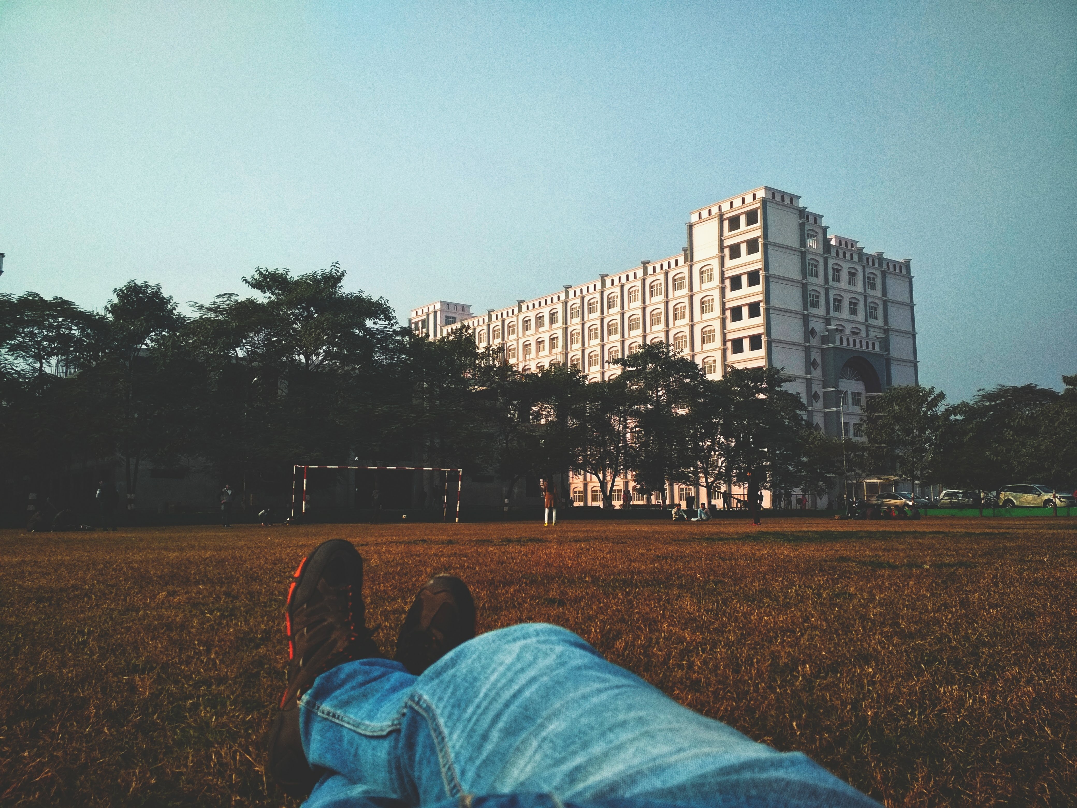 Person in blue denim jean lying on brown grass field looking at white multi-storey building photo