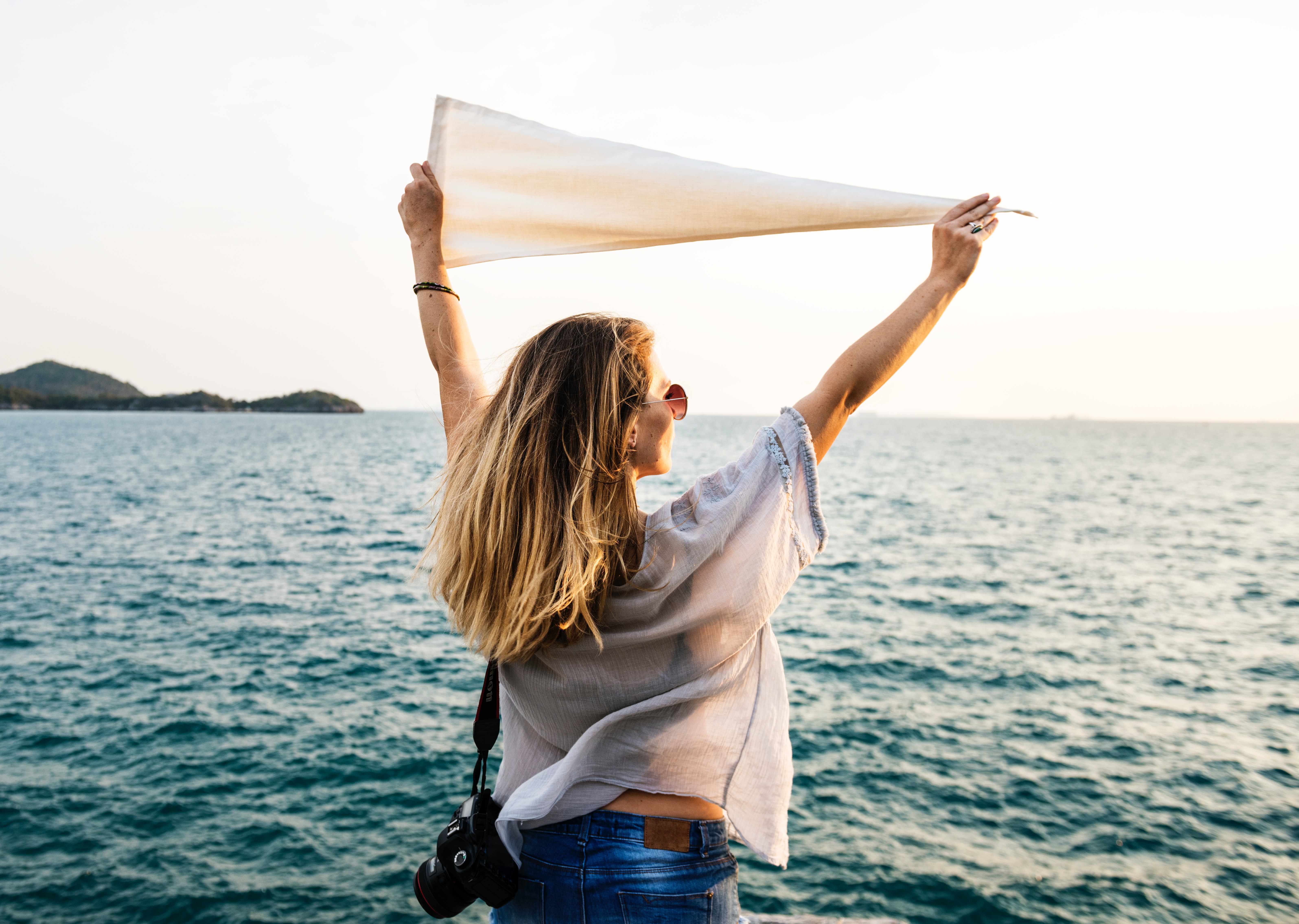 Photo of Woman Wearing White Top, Blue Bottoms and Black Dslr Camera Holding White Textile While Facing the Ocean, Adventure, Tour, Recreation, Relaxation, HQ Photo