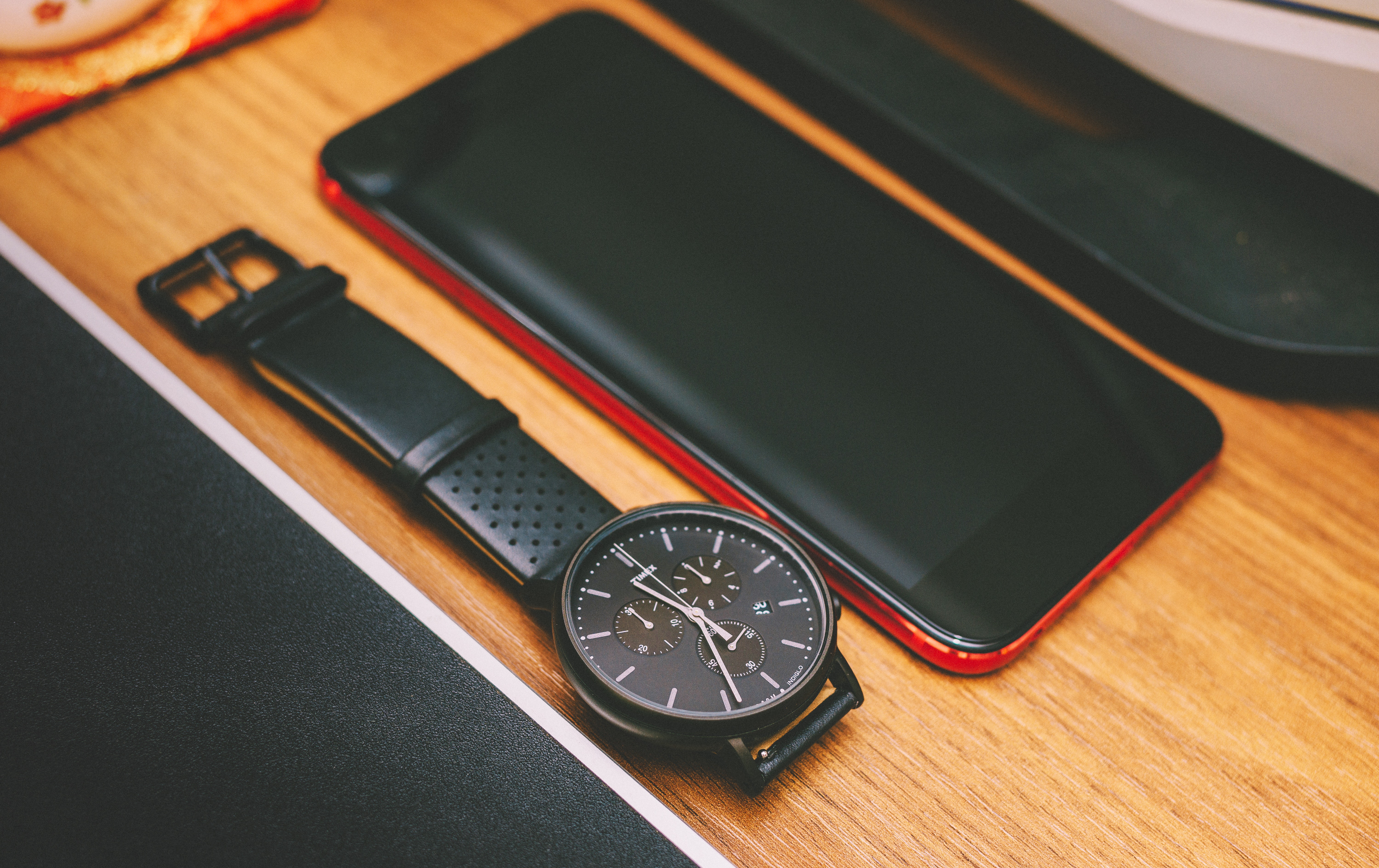 Red smartphone beside the black chronograph watch on brown wooden board photo