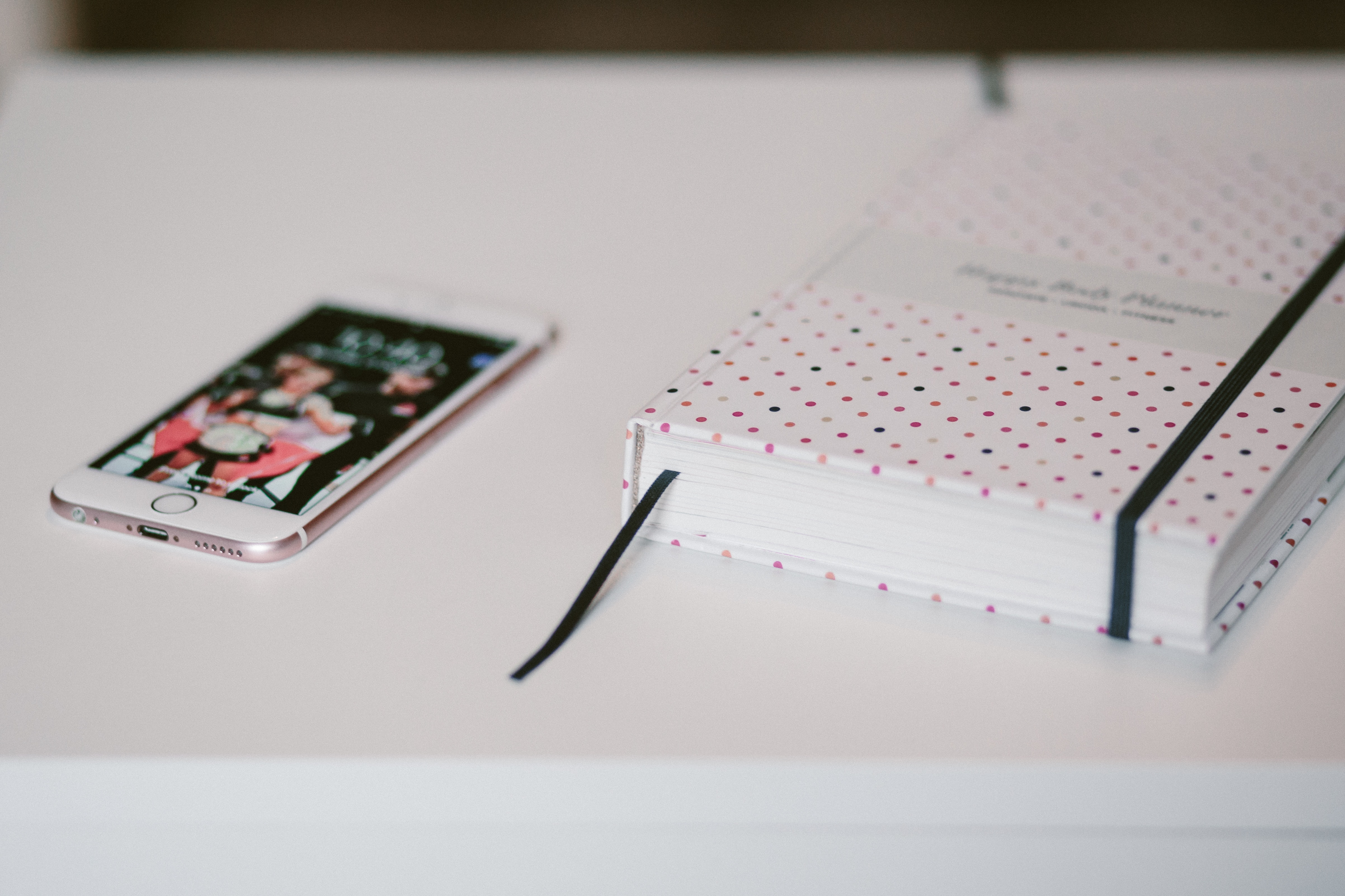 Rose gold iphone 6s beside white and black polka dots book both on top of white wooden table photo
