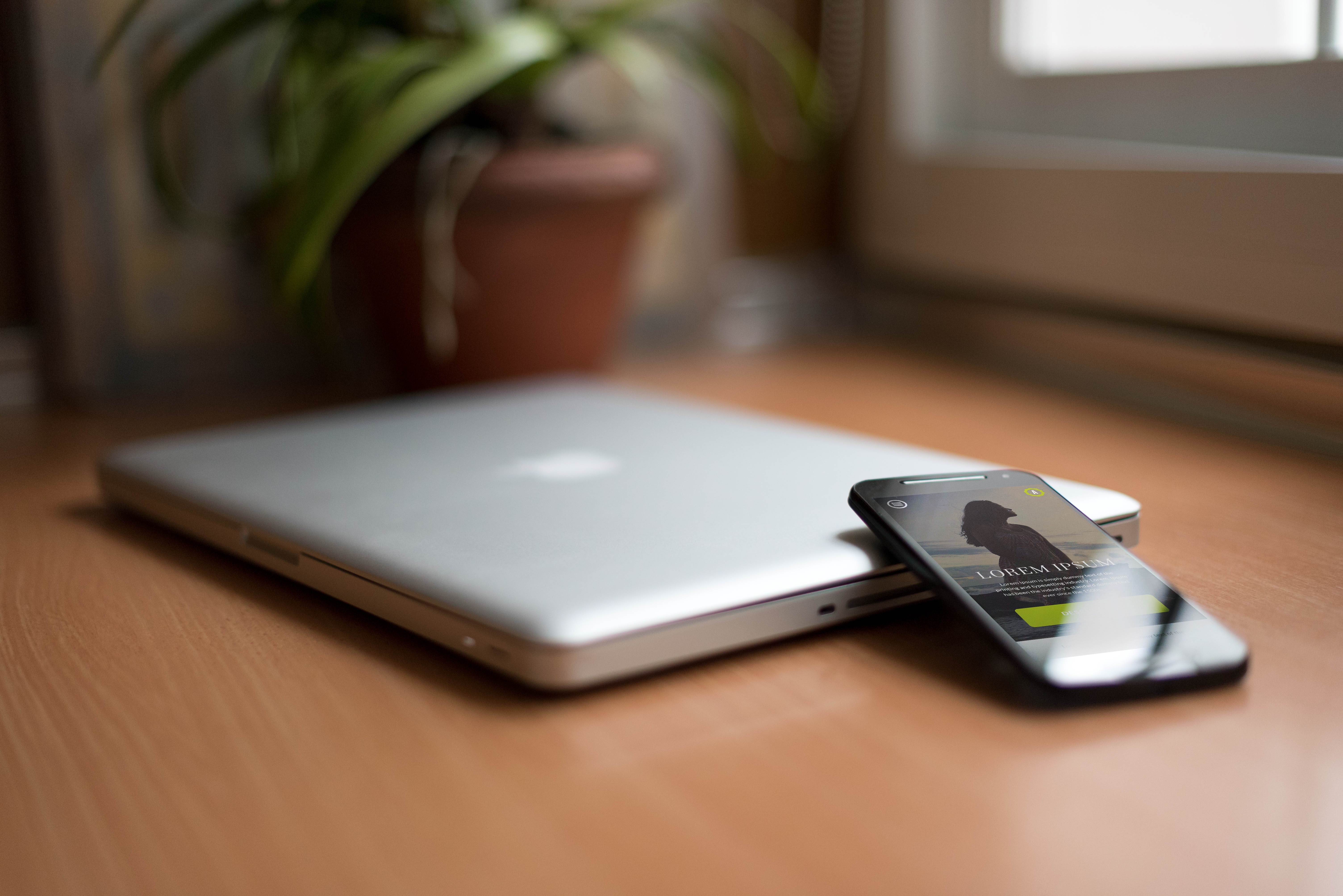 Smartphone beside silver macbook on brown wooden table with potted plant in the background photo