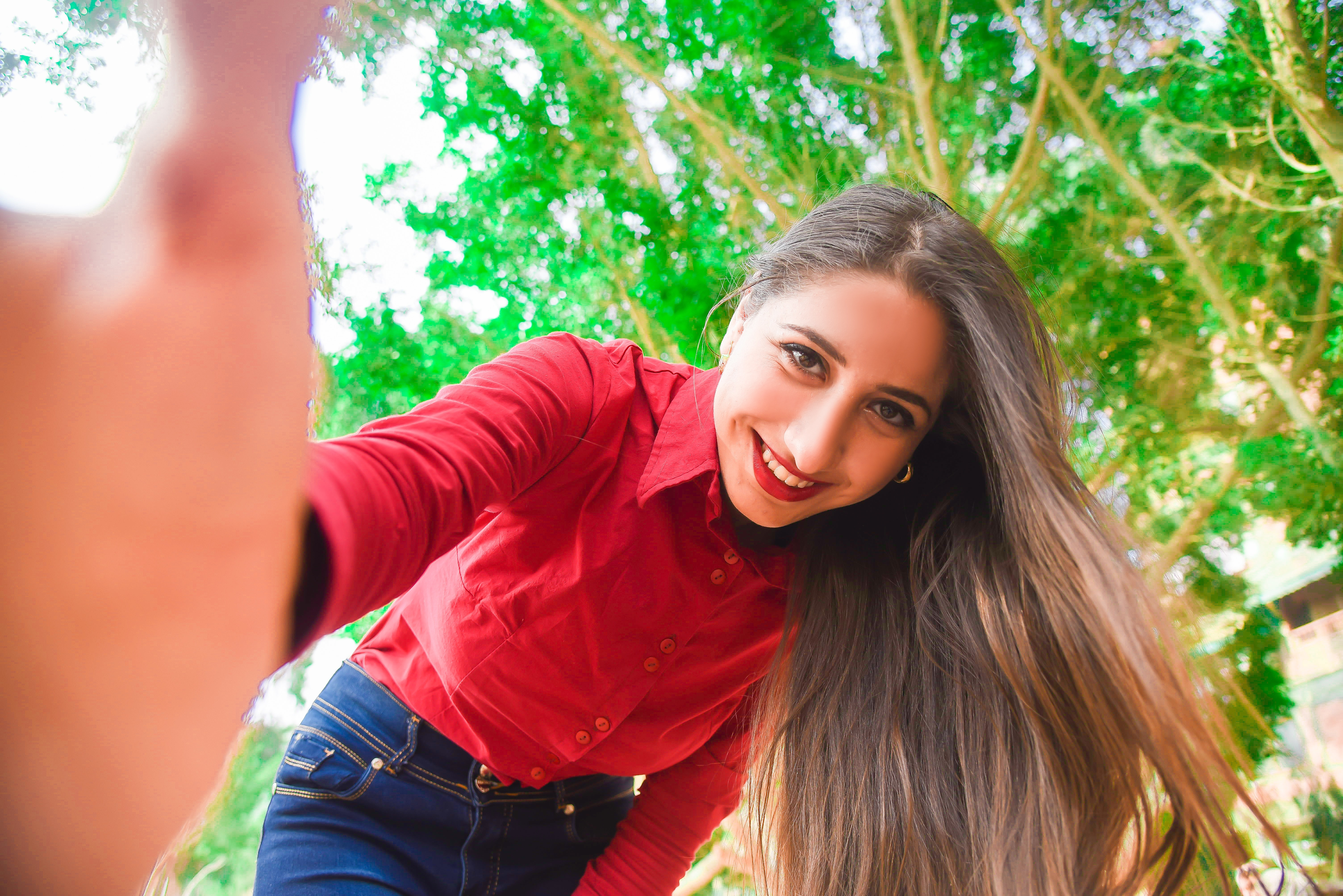 Smiling woman in red shirt and blue jeans taking selfie under green leaved tree photo