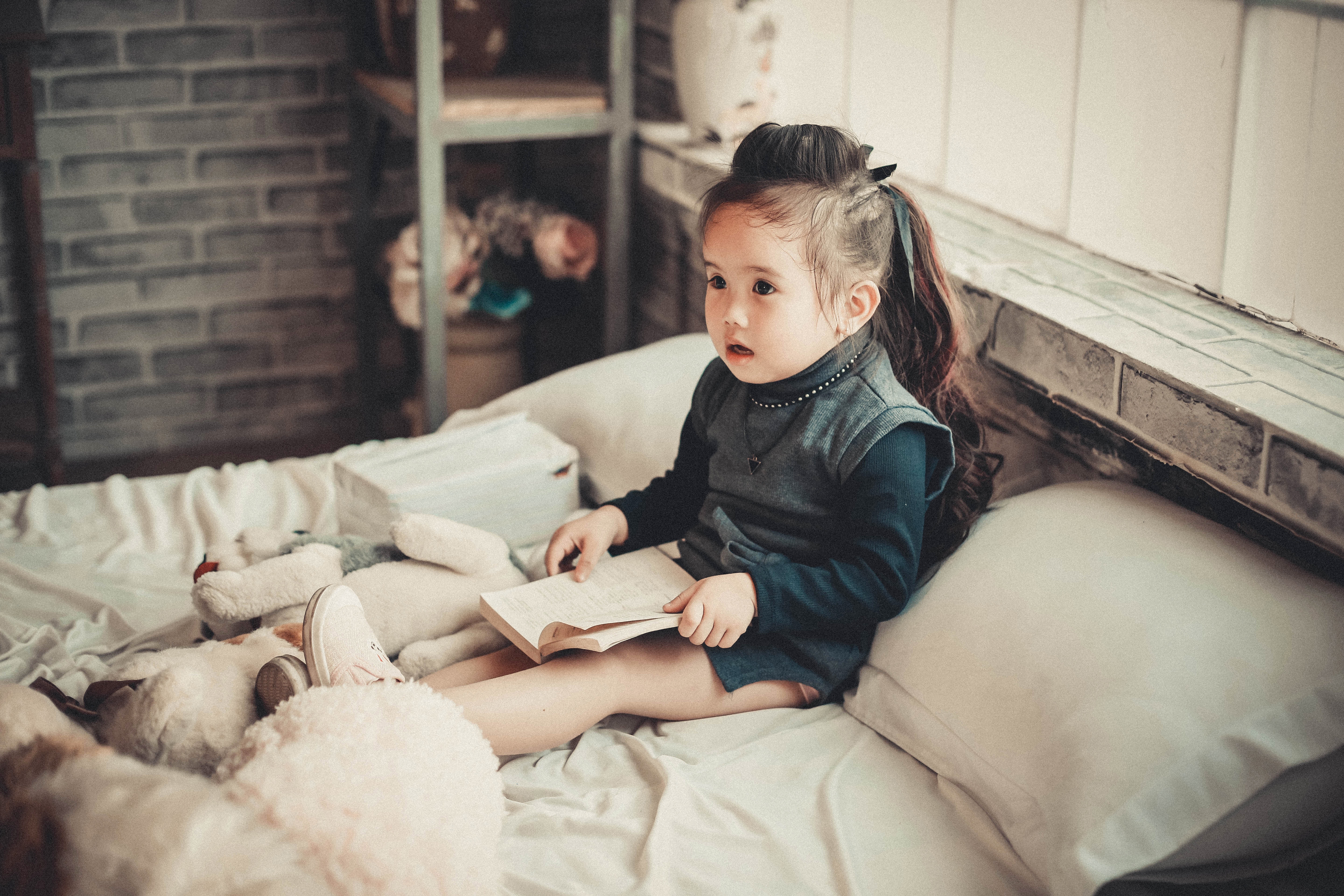 Toddler girl wearing long-sleeved top reading book while sitting on bed photo