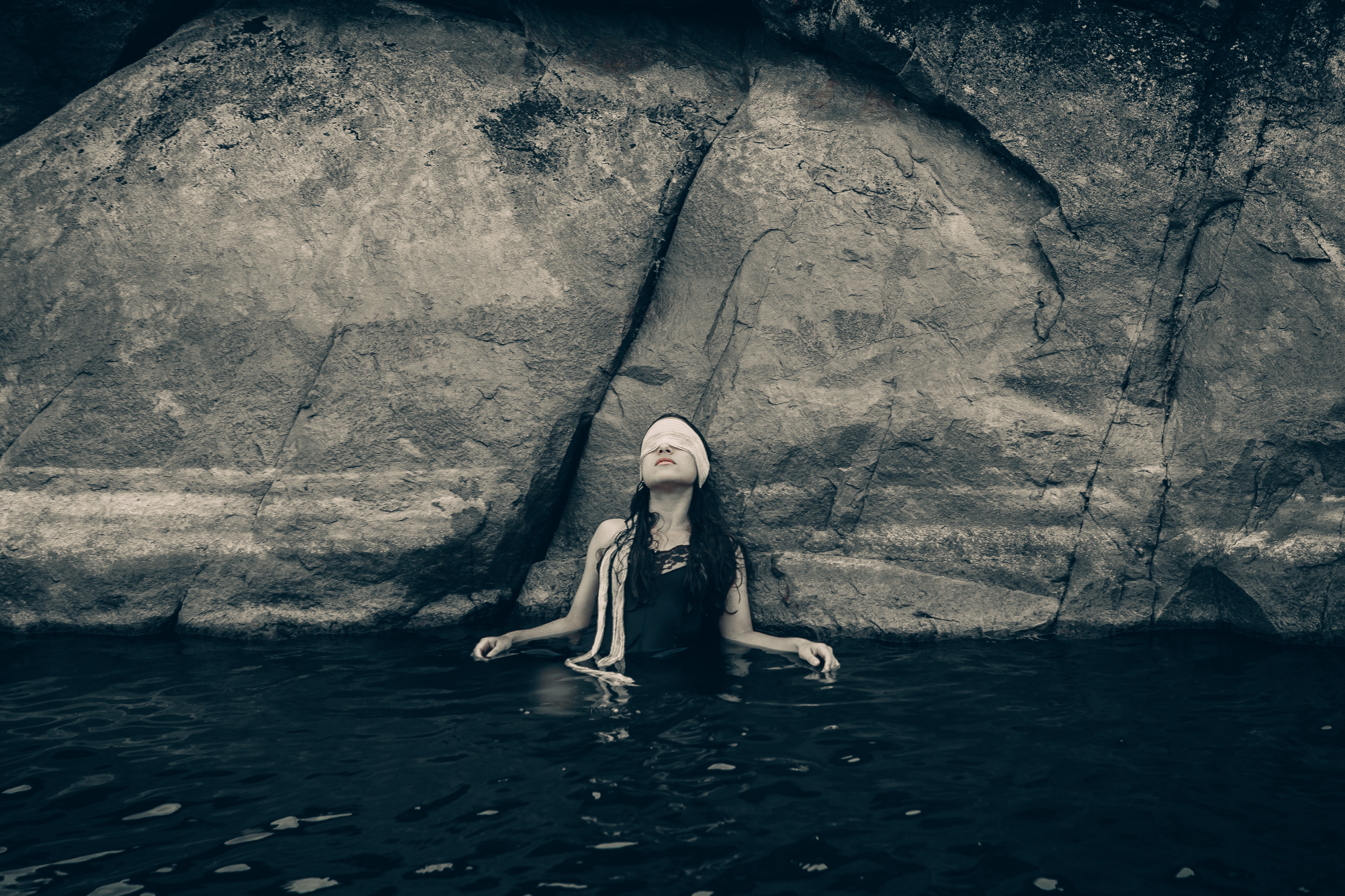 Woman in blindfold wearing black top on body of water while leaning on a rock photo