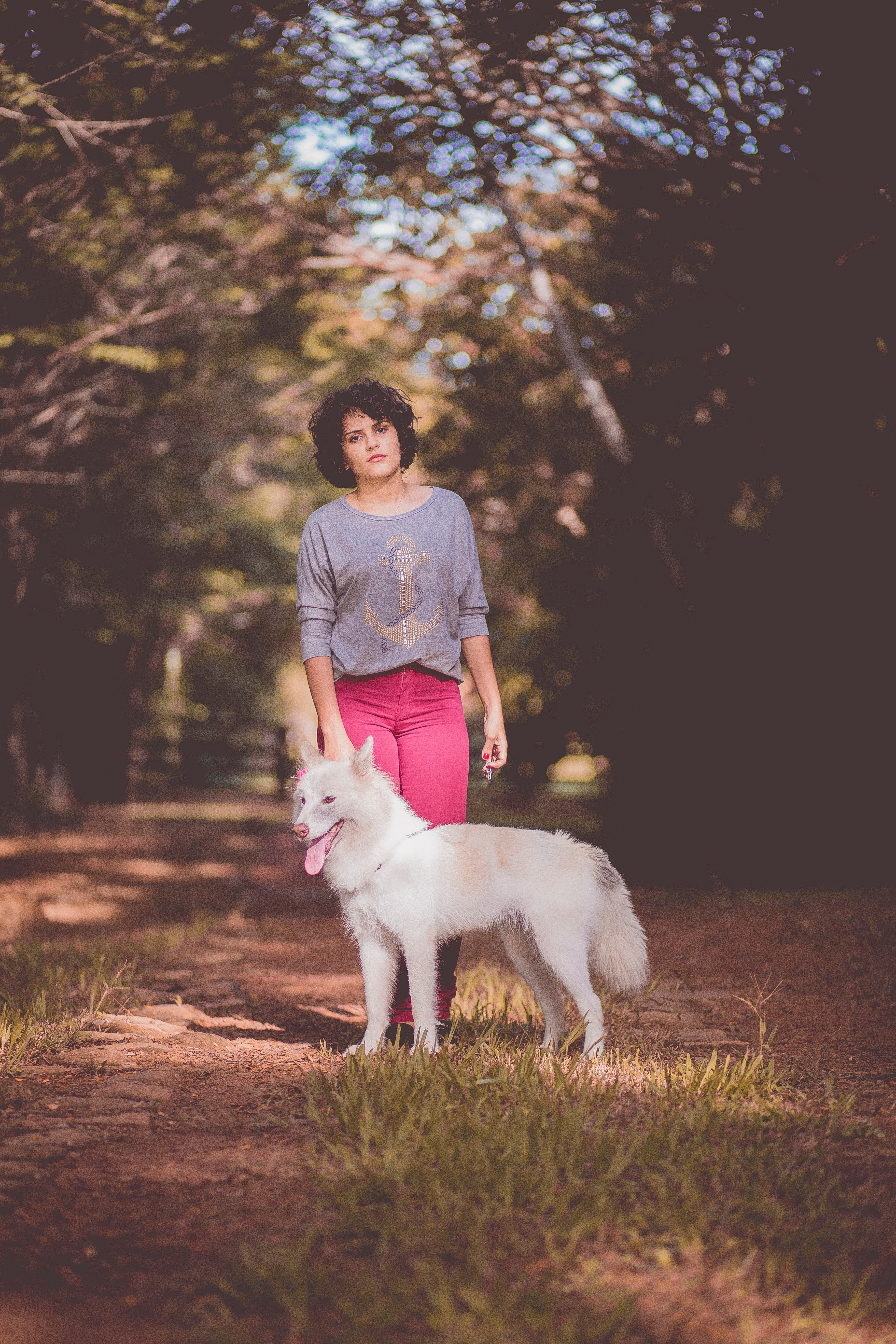 Woman in purple long sleeve shirt and pink jeans standing next to white german spitz with background of green trees during day photo