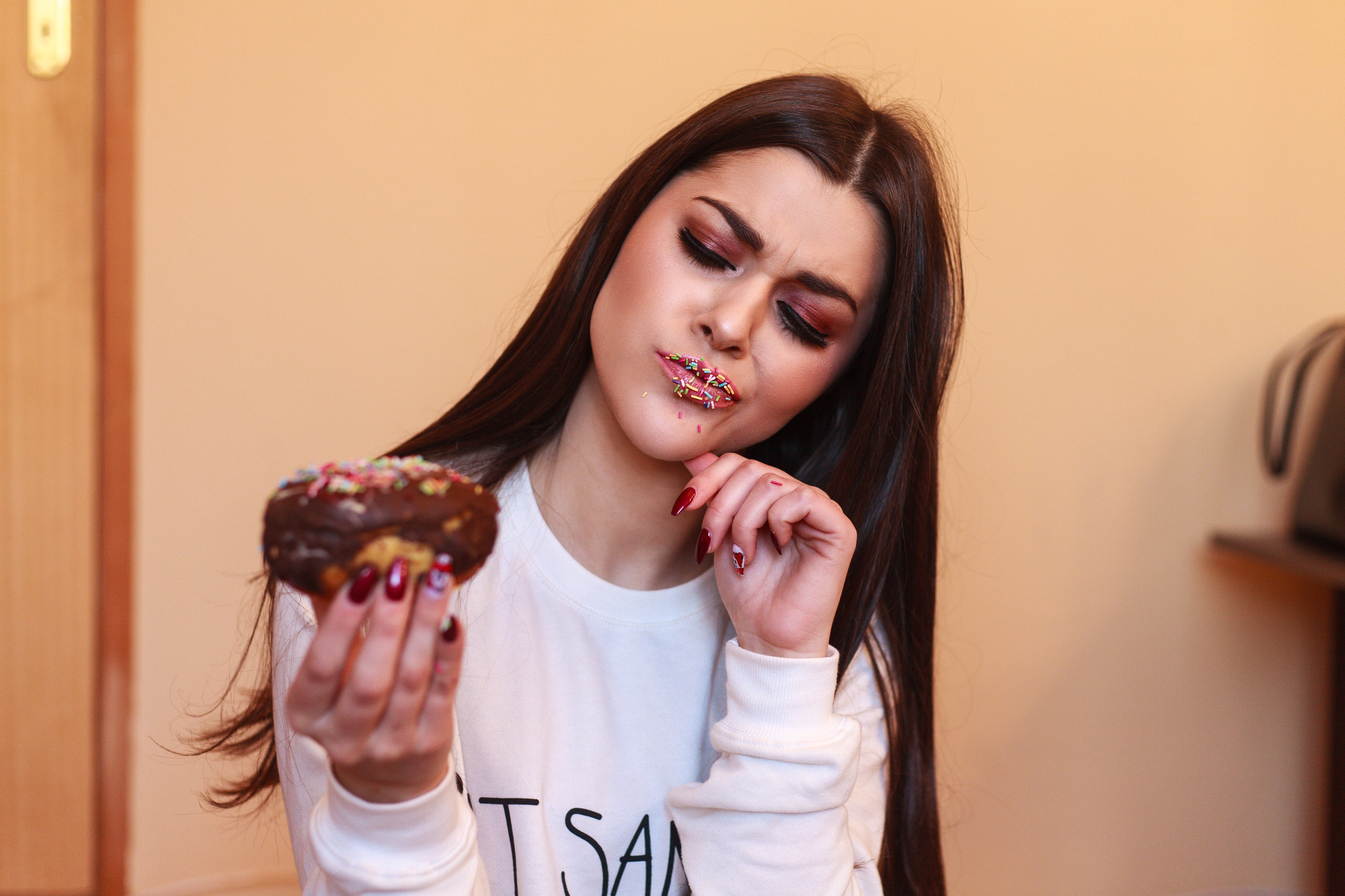 Woman in white crew-neck sweatshirt holding and looking at chocolate doughnut inside room photo