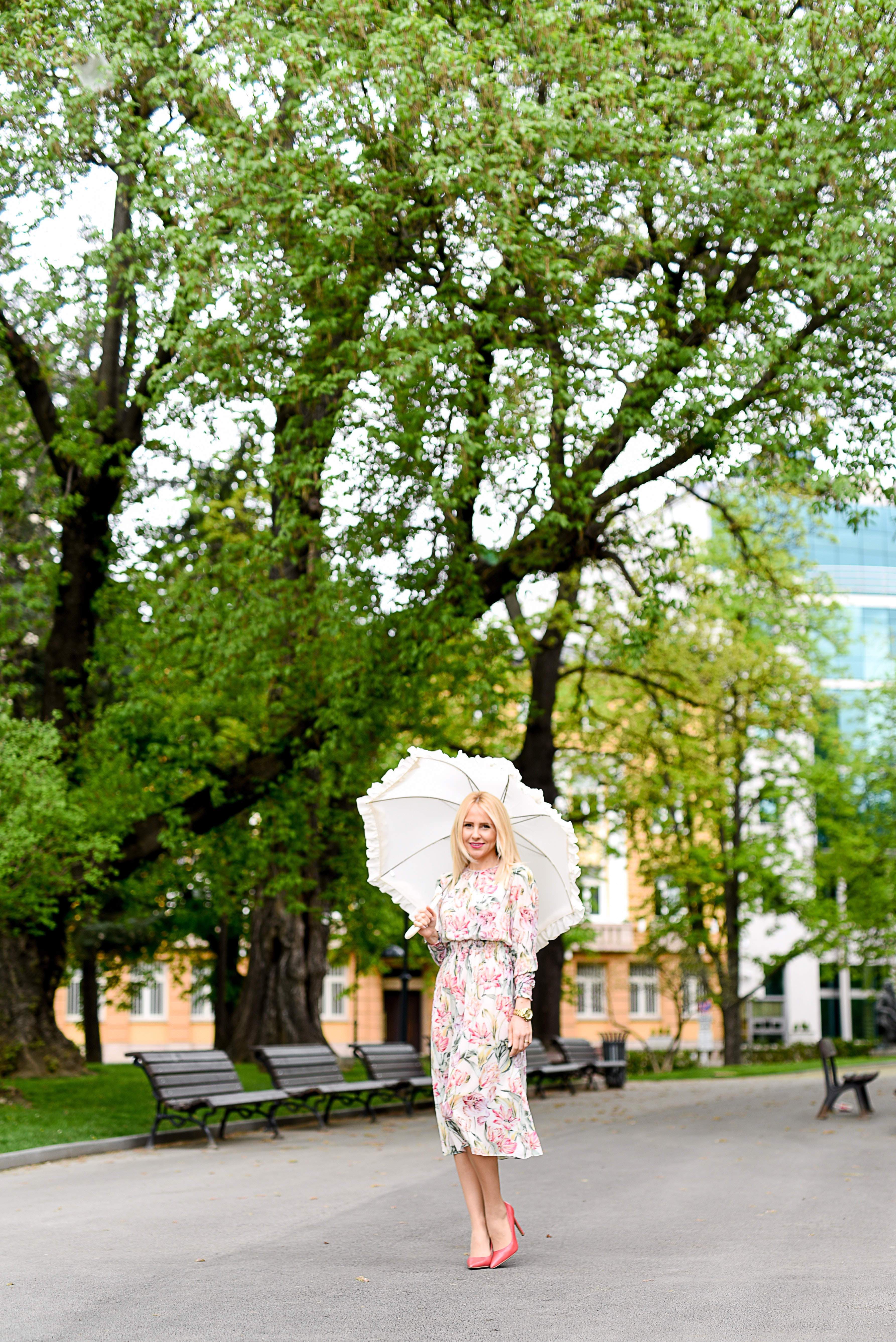 Woman in white green and red floral holding white umbrella on gray concrete pathway near brown wooden bench near trees during daytime photo