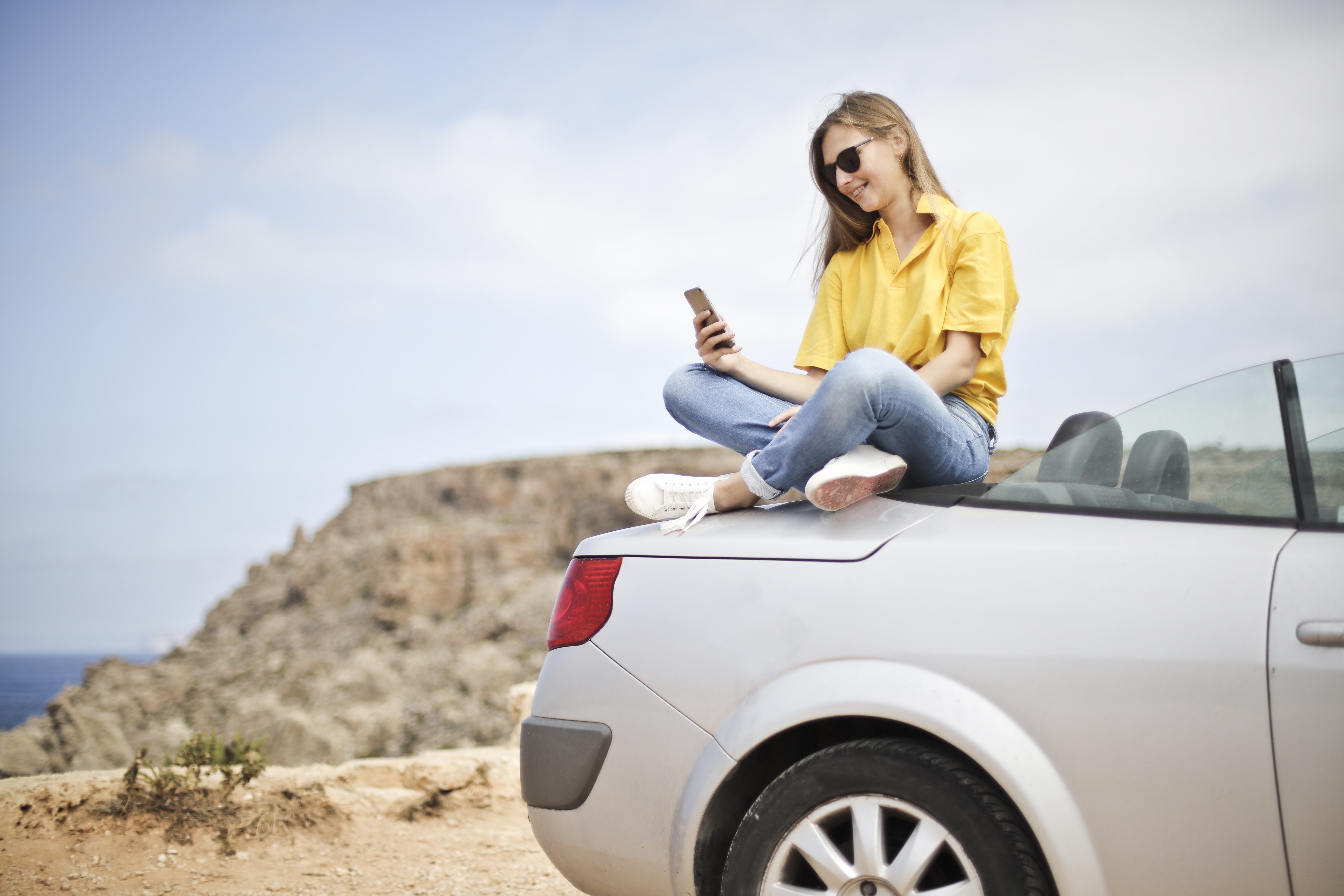 Woman in yellow blouse and blue jeans taking selfie while sitting on car photo