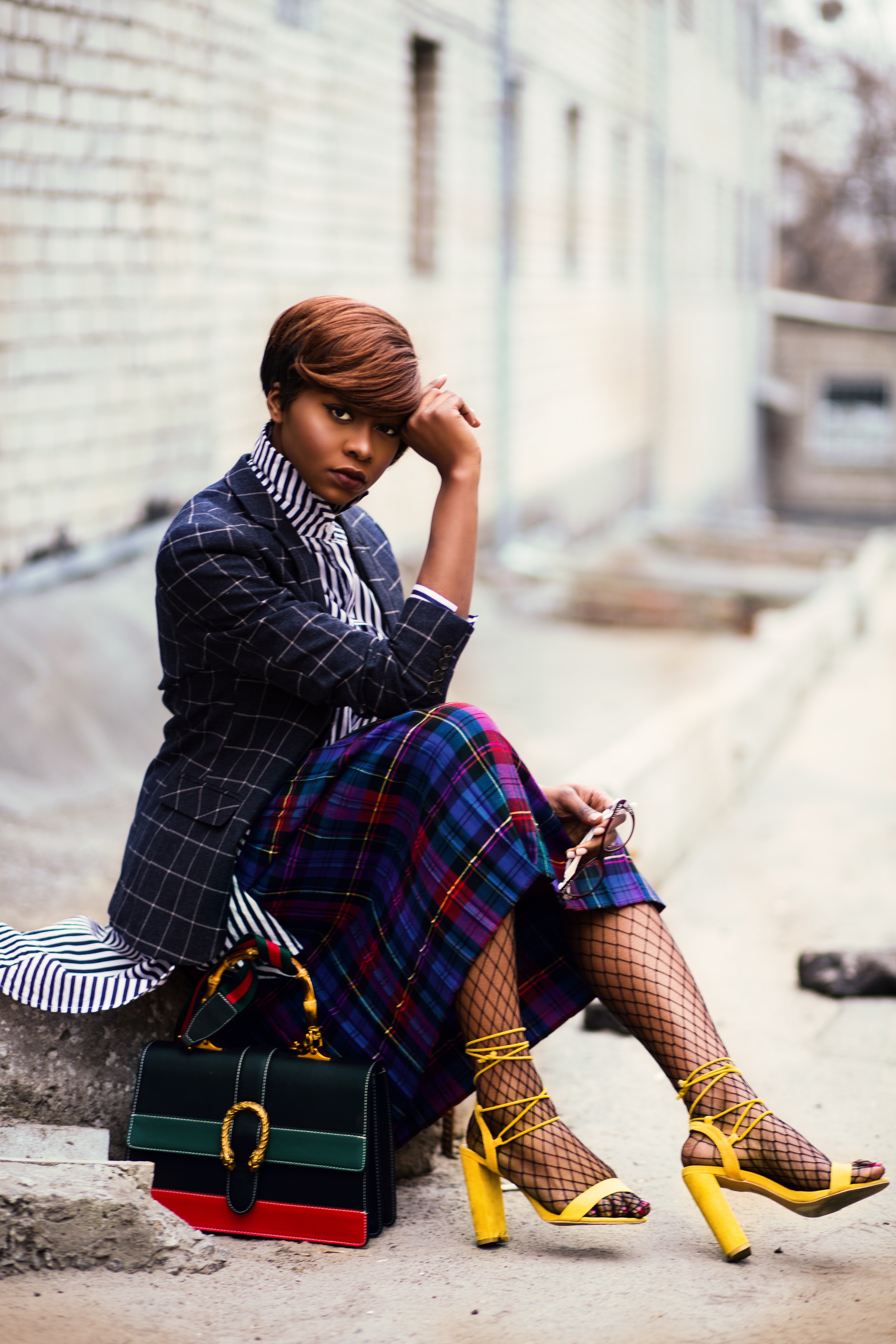 Woman wearing black and grey tattersall blazer and multicolored plaid skirt with black mesh stocking and yellow chunky heeled sandals sitting on grey concrete pathway photo