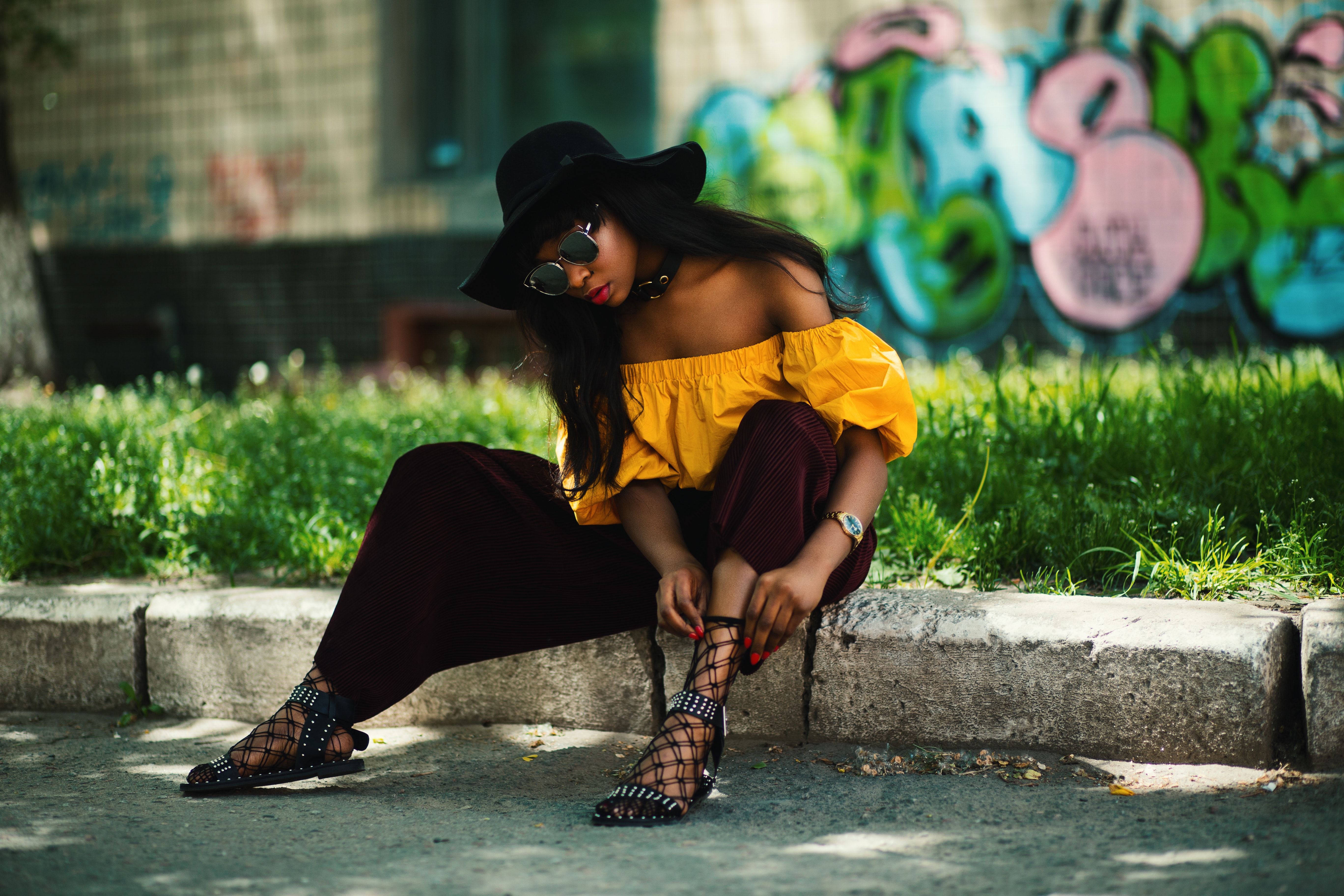 Woman wearing yellow off-shoulder top and black pants sitting on sidewalk fixing lace sandals photo