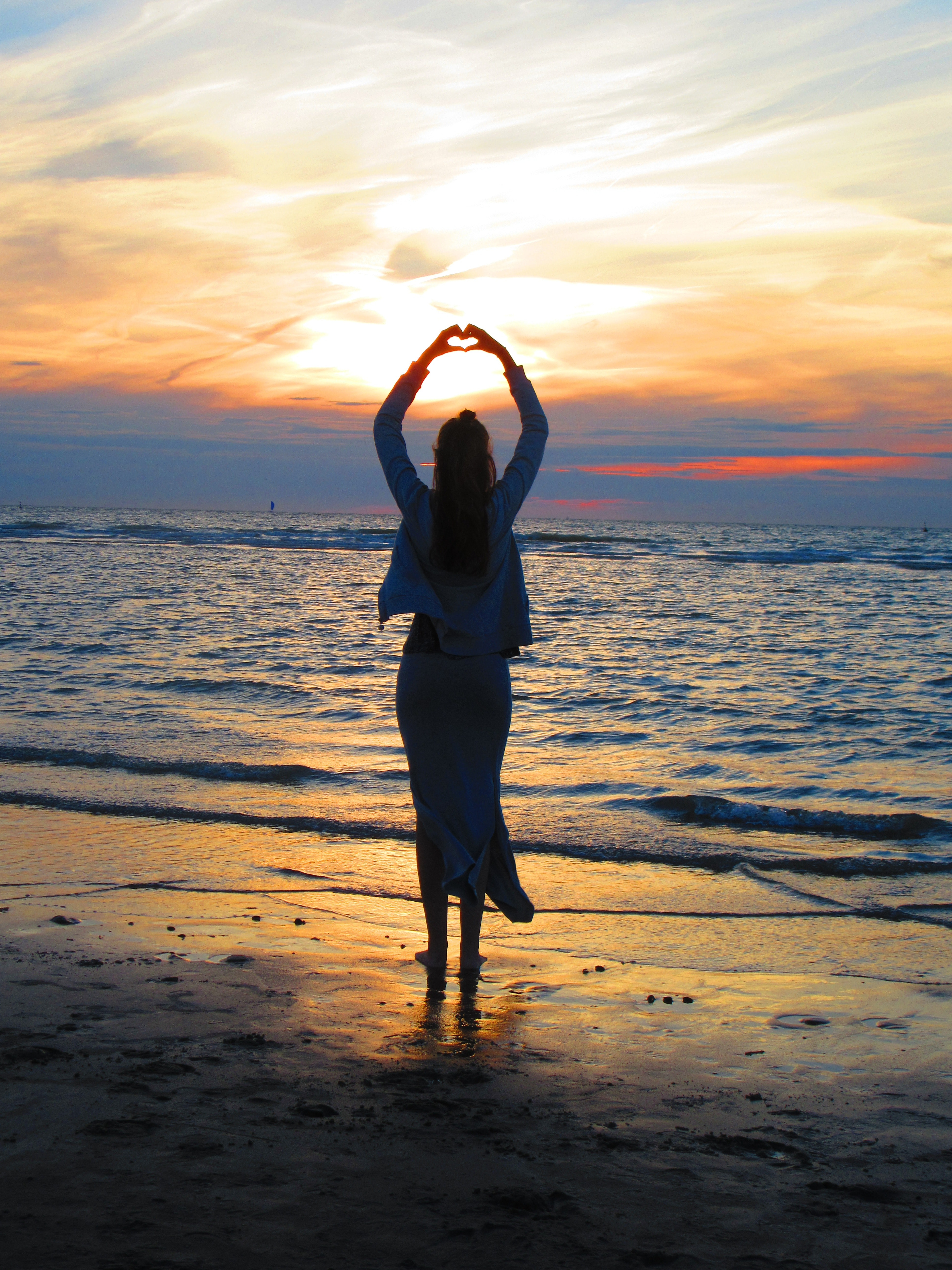 Woman with arms up making heart sign while standing on beach at sunset photo