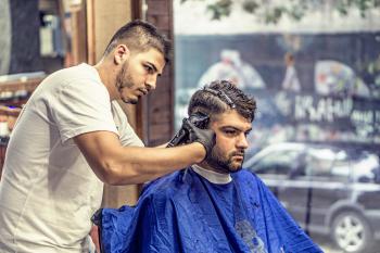 Barber in White Shirt Trimming Man's Hair in Blue Textile While Sitting Nearby Glass Window