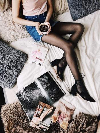 Flatlay Photography of a Woman Holding White Mug With Black Liquid While Lying on a Bed Surrounded by Fur Pillows and Magazines