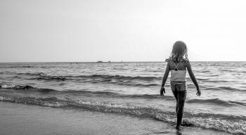 Grayscale Photo of Girl Walking on Seashore With White Spaghetti Strap Top