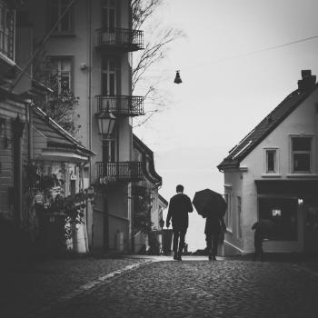 Grayscale Photo Of Man Beside Woman Under Umbrella Walking On Pavement