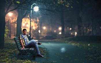 Man in Blue Denim Jeans Sitting Down by Wooden Bench Near Post Lamp Lighted