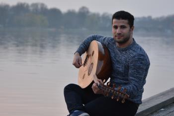 Man in Gray Crew Neck Sweatshirt and Black Pants Sitting in Gray Concrete Holding String Instrument Near Body of Water