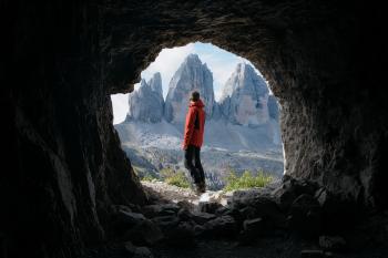Man in Red Jacket Standing Outside of Cave in Front of Three Mountains