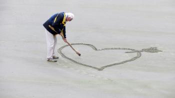 Man Wearing Blue Jacket Holding a Brown Stick Towards the Heart Drawn on Sand