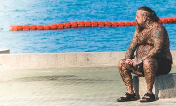 Man With Tattoos Sitting on Gray Concrete Floor Near Body of Water