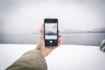 Photo of Person Wearing Brown Coat Holding Android Smartphone While Taking Picture of Mountain and Body of Water