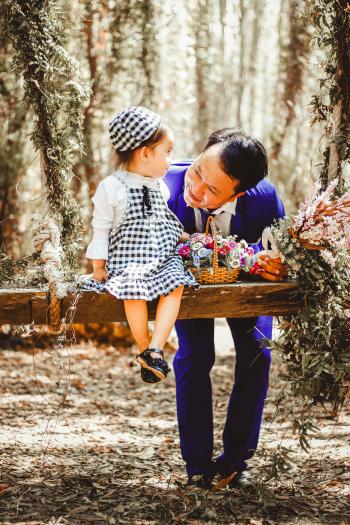 Selective Focus and Color Photography of Man Looking at Her Girl Sitting on Garden Swing White Holding Bouquet of Flower in Brown Wicker Basket