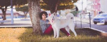Woman in Gray Long Sleeve Top and Red Pants Sitting Beside Tree and White Medium Coated Dog