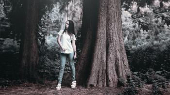 Woman in T Shirt Posing Beside Rough Bark Tall Tree in Grayscale Photography