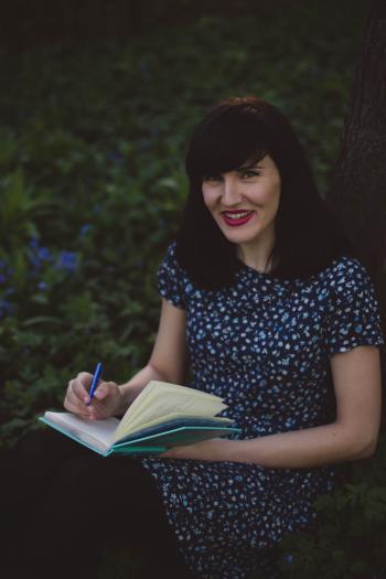Woman in White and Blue Floral Crew Neck Short Sleeve Shirt Holding a Teal Notebook Leaning in Brown Tree Trunks