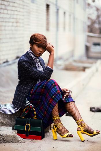 Woman Wearing Black and Grey Tattersall Blazer and Multicolored Plaid Skirt With Black Mesh Stocking and Yellow Chunky Heeled Sandals Sitting on Grey Concrete Pathway