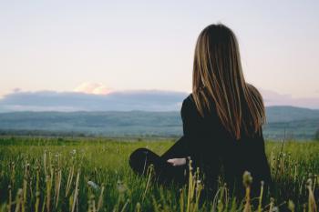 Woman Wearing Black Long Sleeved Shirt Sitting on Green Grass Field Near Mountain Under Cloudy Sky