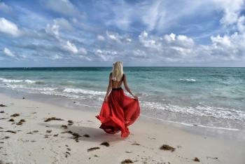 Woman Wearing Red Dress Walking on Seashore Under Blue and White Sky