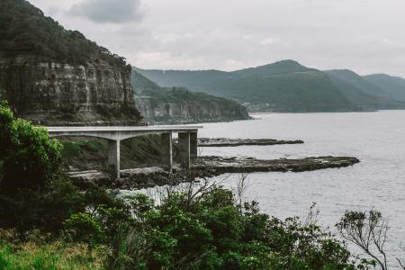 Concrete Bridge Near Mountain Above Shoreline Under Cloudy Sky