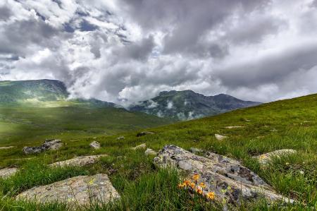 Green Grass Field With Rocks Near Mountains during Cloudy Daytime Sky